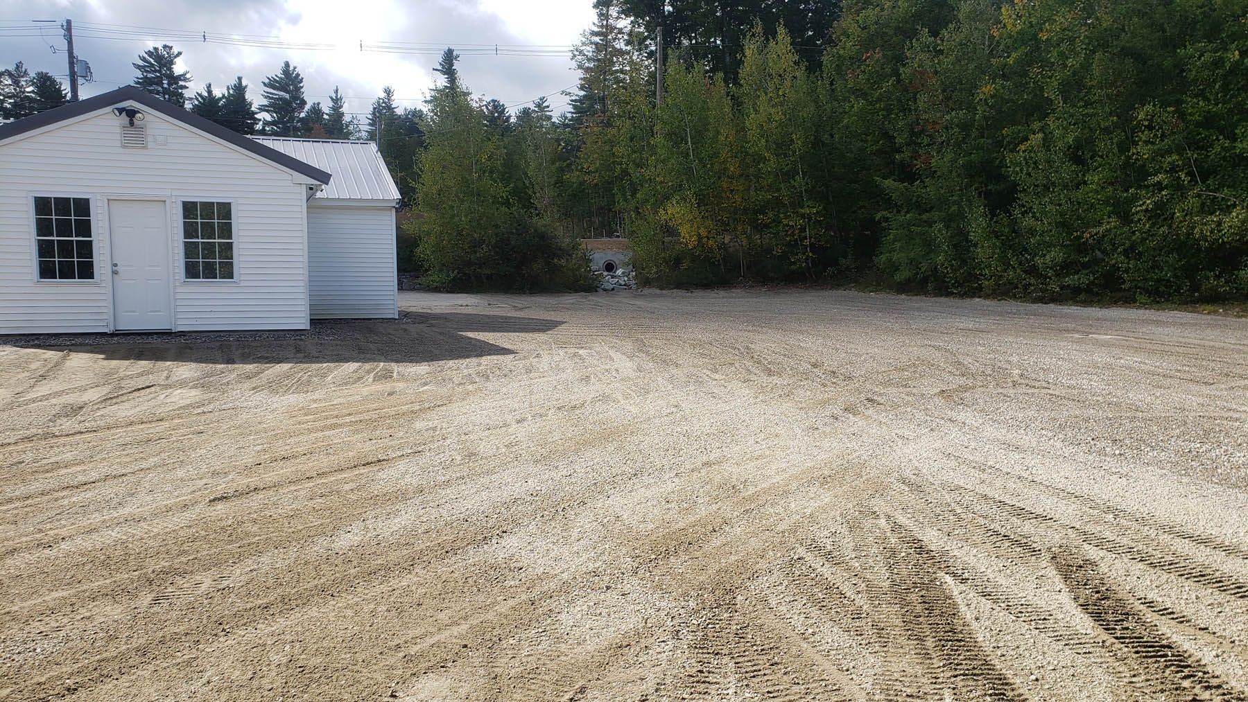 White shed on a gravel lot, flanked by green trees, under a cloudy sky.