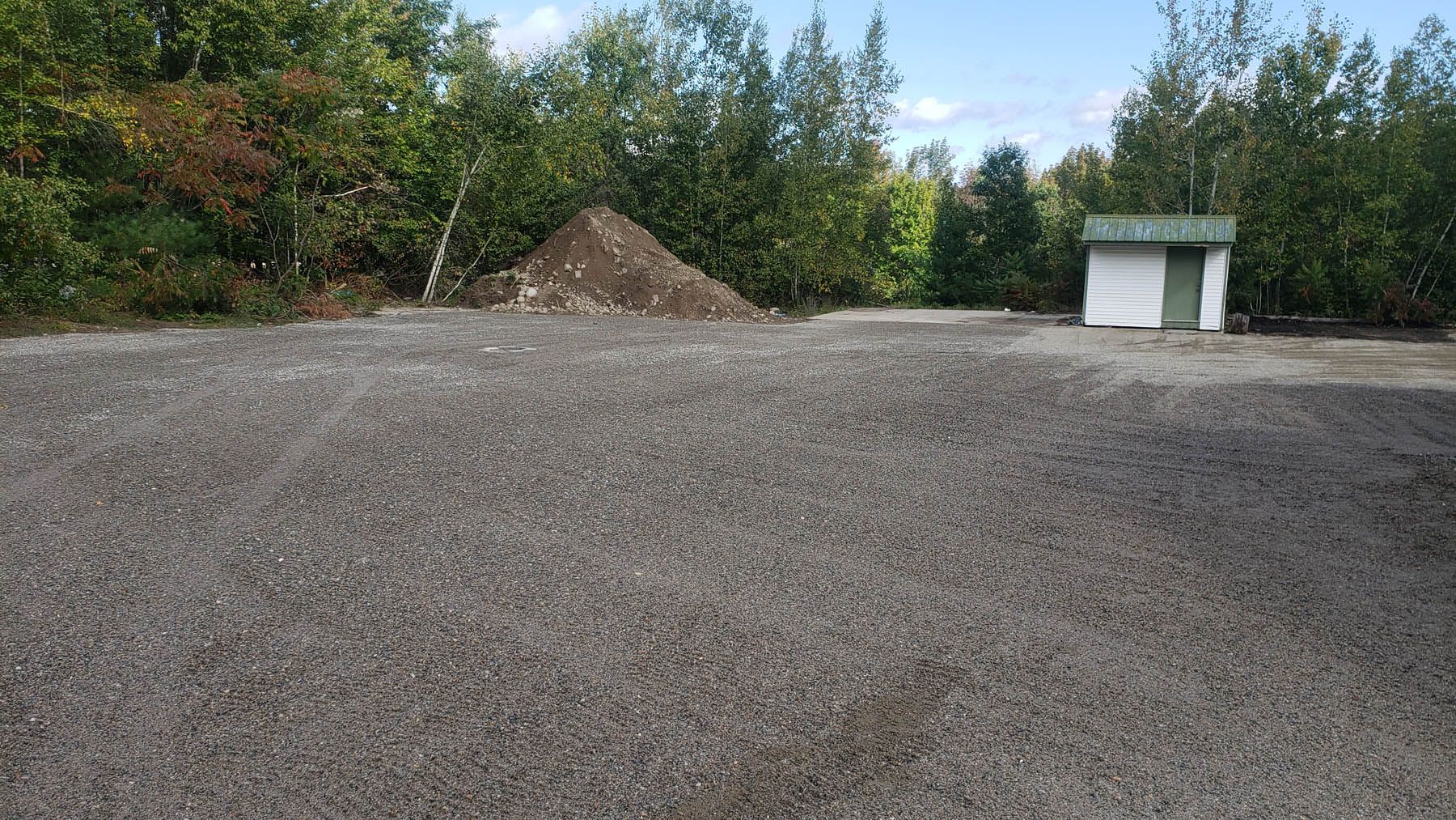 Gravel lot with a dirt pile, small shed, and trees in the background.