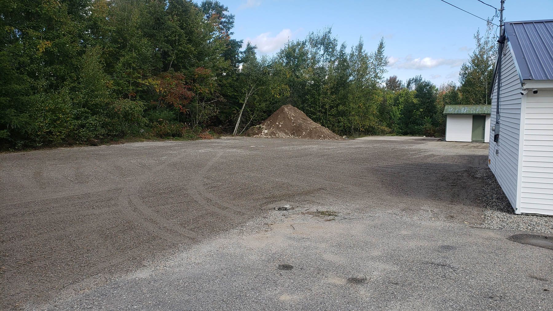Gravel lot next to a white building and trees. A pile of dirt sits further back. Blue sky.