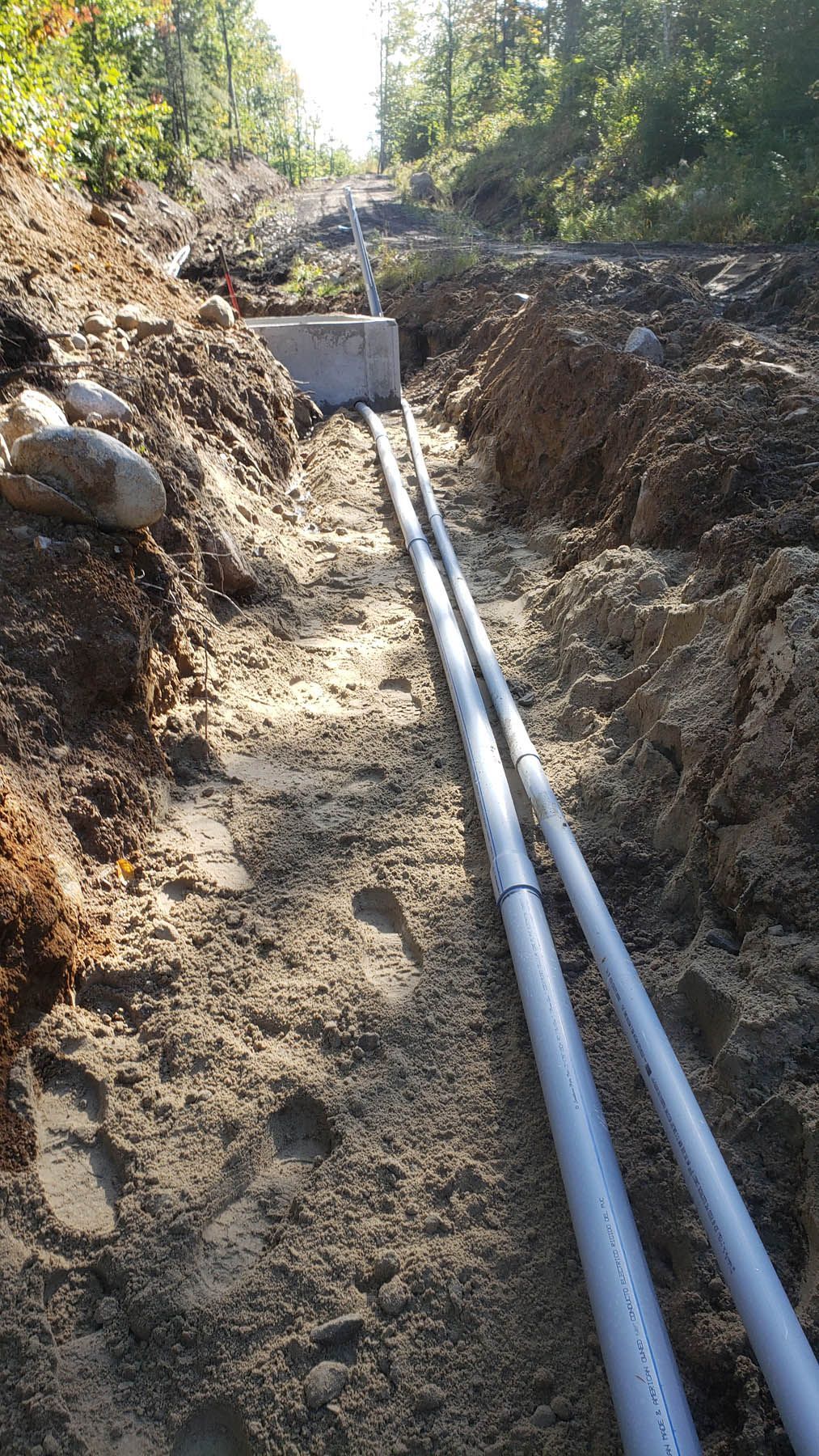 Two gray pipes run through a trench dug in dirt, heading toward a concrete block on a hillside.