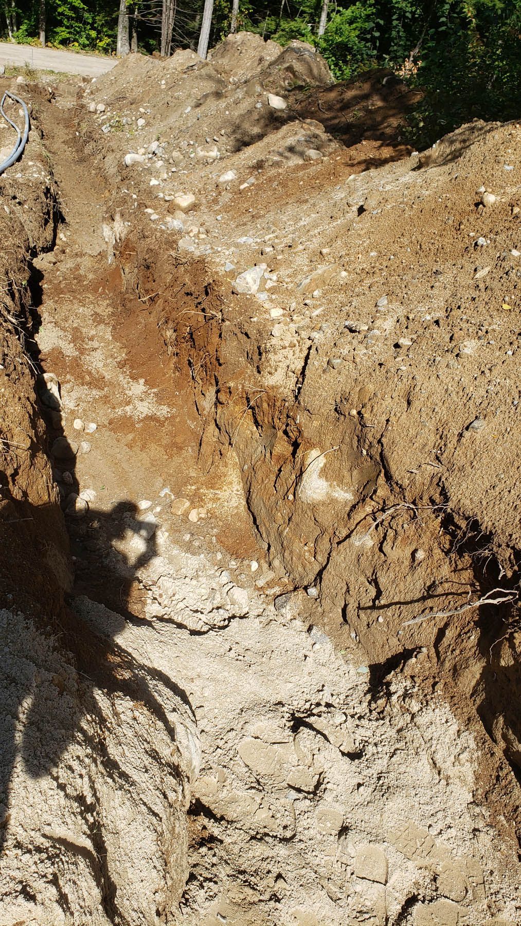 A narrow trench dug in sandy, gravelly soil, with mounds of excavated earth alongside.