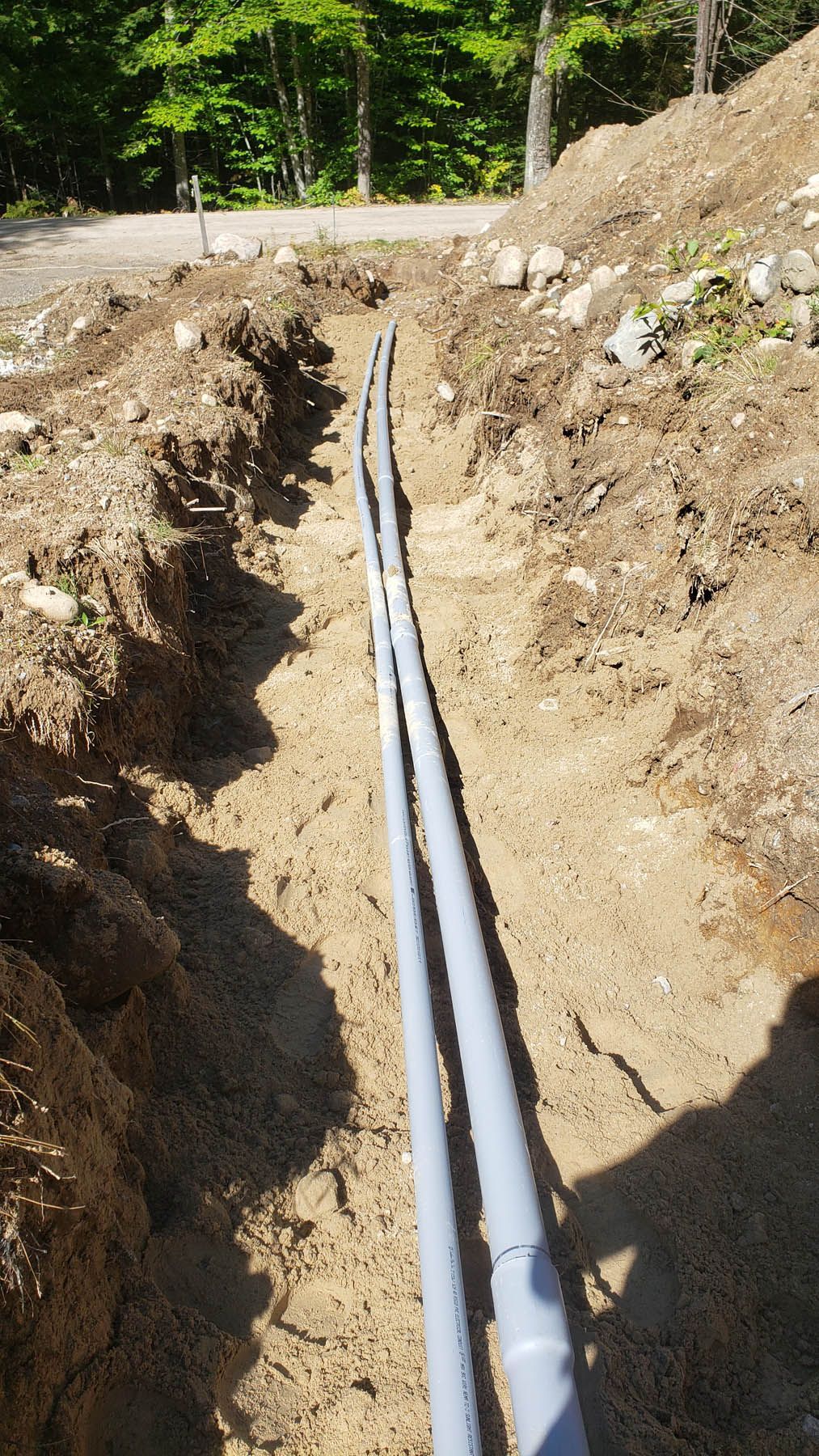 Two gray pipes running through a narrow dirt trench, in a forested area.