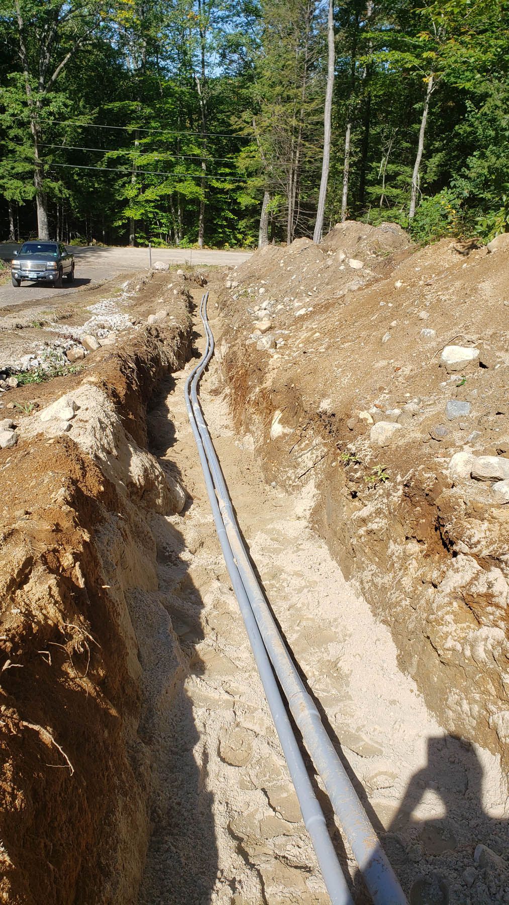 A trench holds two gray pipes, with gravel at the bottom. The scene is outdoors in a dirt area.
