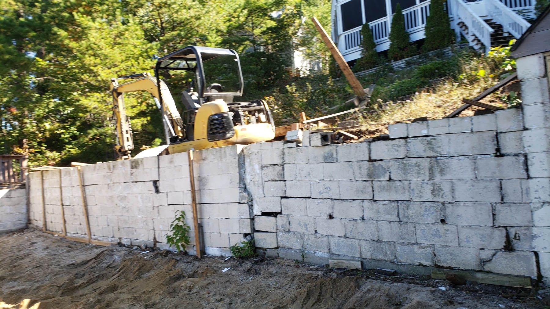 Mini excavator on a concrete block retaining wall. A house is visible on a hillside in the background.