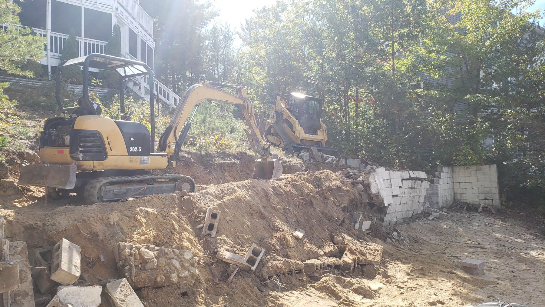 Two yellow excavators demolish a concrete wall on a hillside, with a house in the background.
