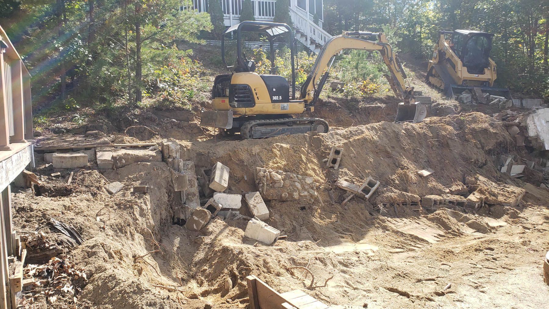 Two excavators working on a hillside with a house in the background; dirt and construction debris.