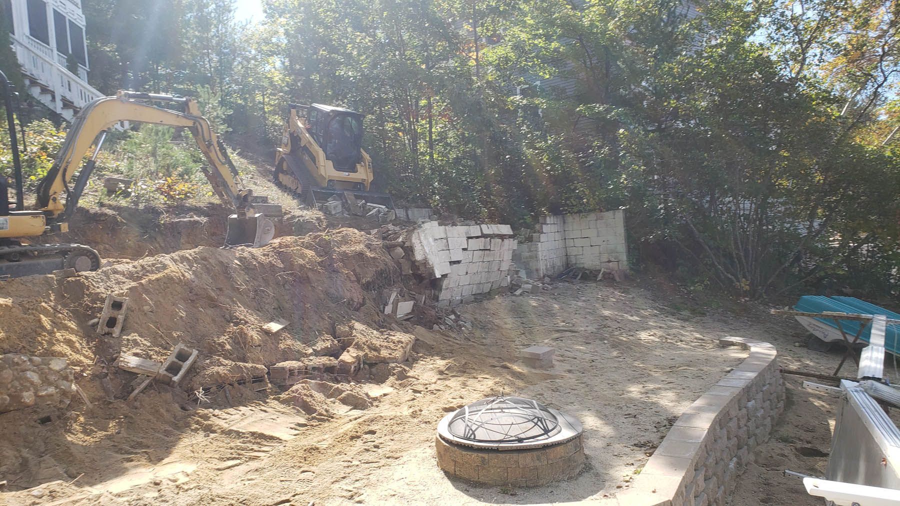 Construction site with excavator removing dirt and debris next to a white structure and trees.