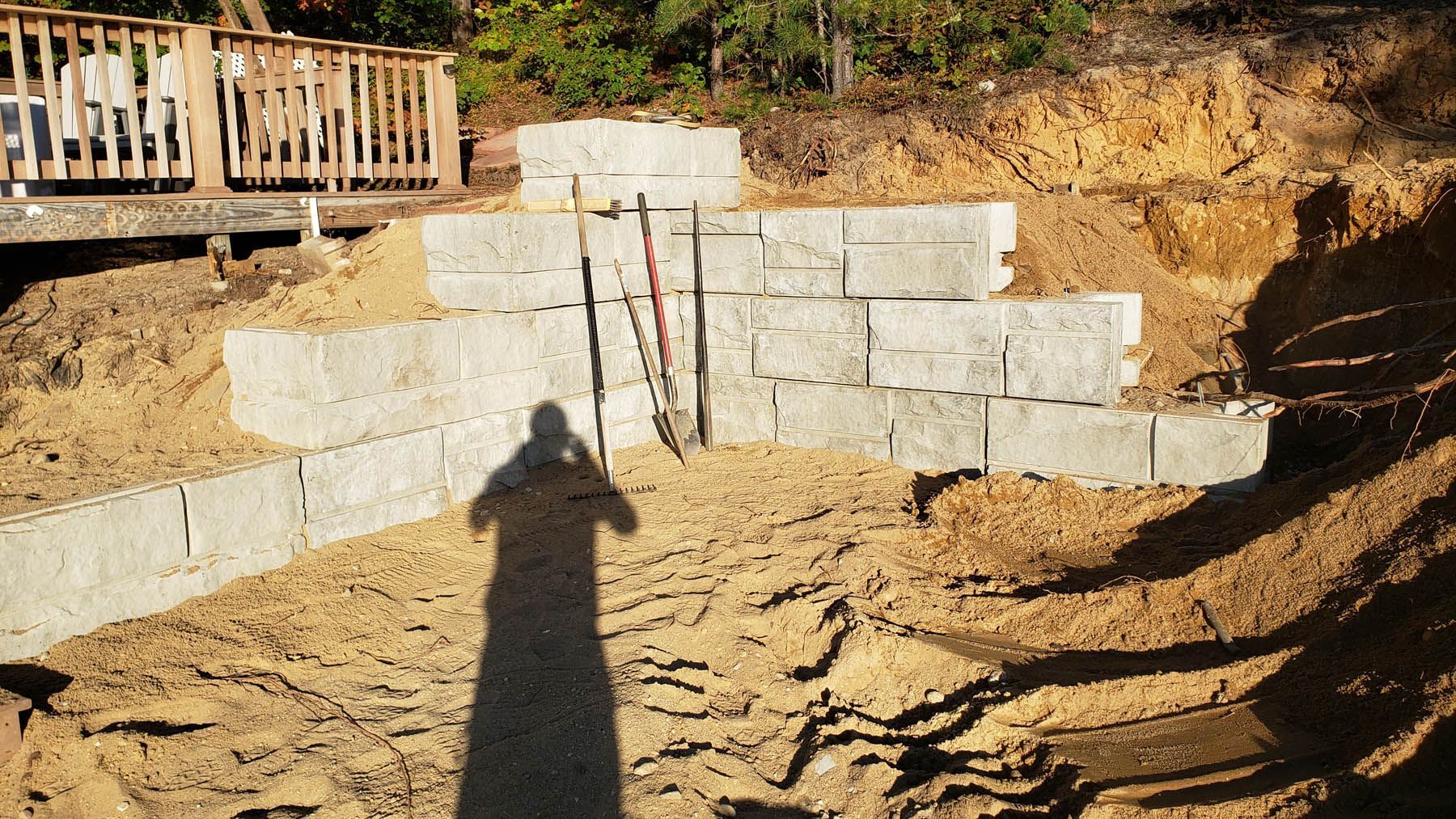 Concrete block retaining wall under construction with sand and tools, person's shadow.