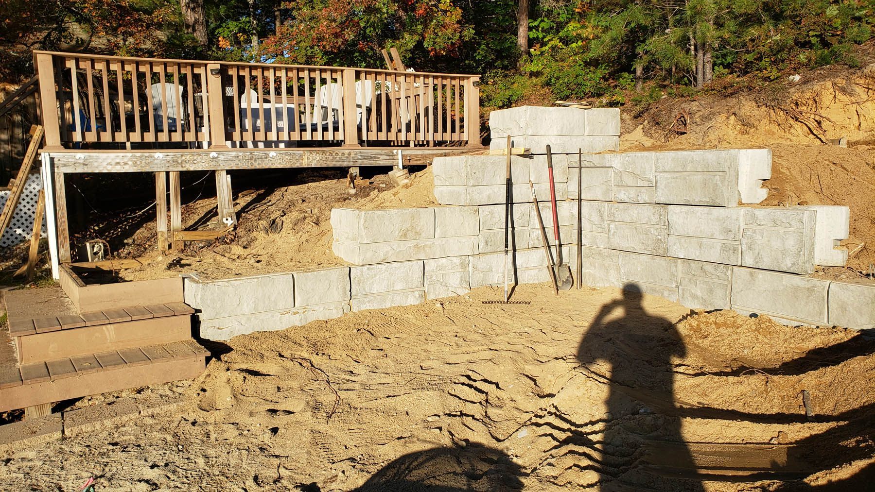 A tiered retaining wall under construction near a deck with tools. Brown dirt, gray blocks, and a shadow.