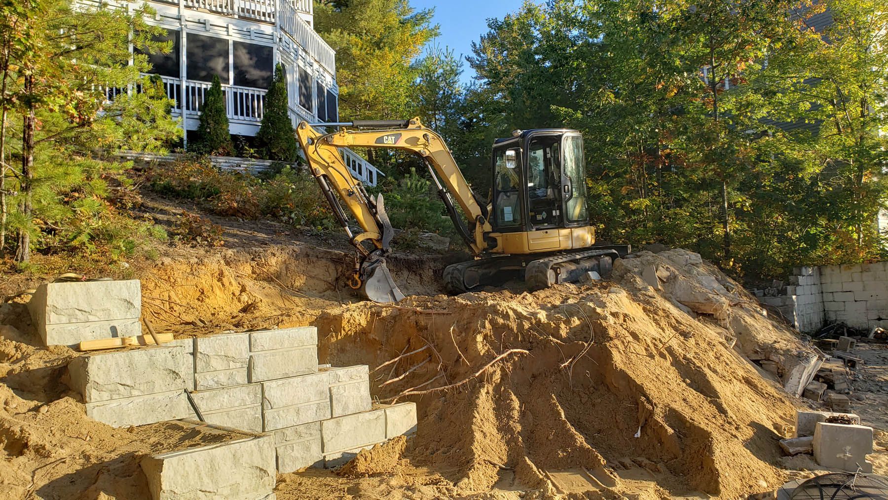 Excavator on a sandy hillside, building steps with concrete blocks, near a house with a porch.