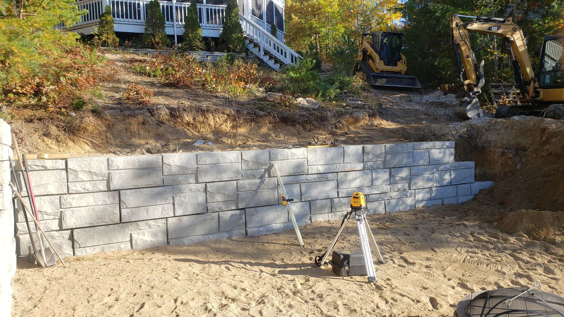 Retaining wall construction at a residential property. Gray blocks, a yellow excavator, and a laser level are visible.