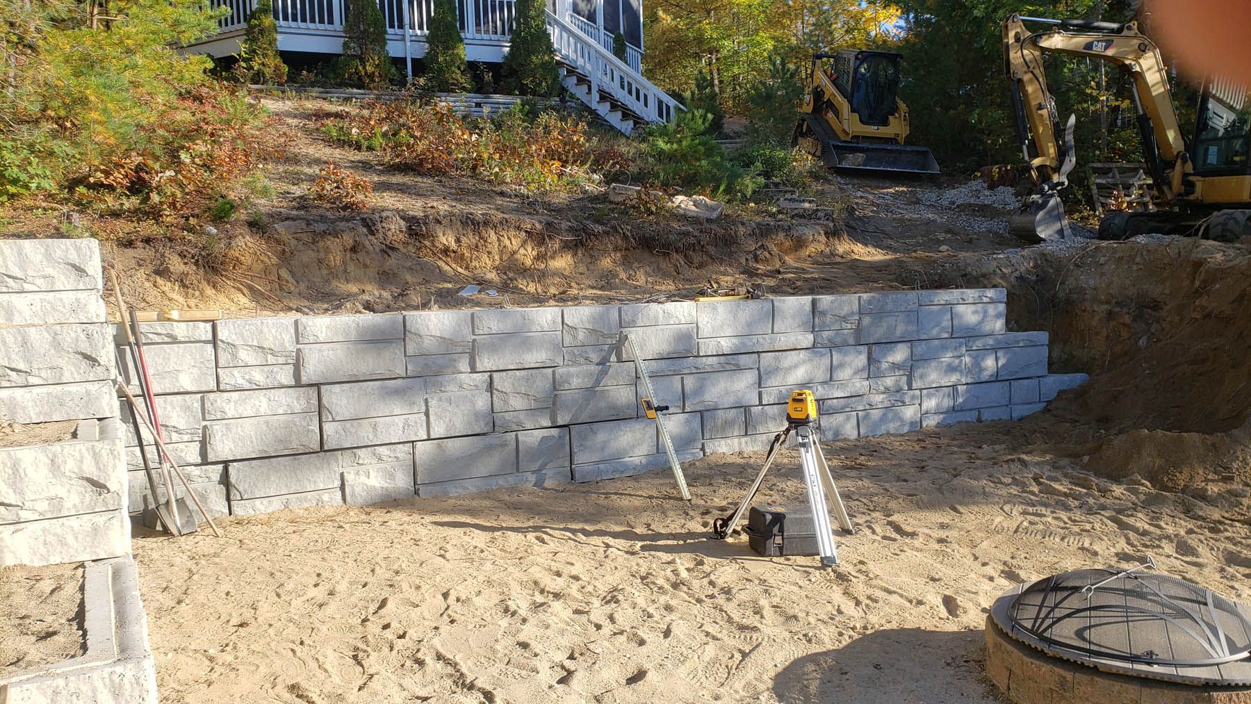 Granite block retaining wall under construction on a hillside; an excavator is visible in the background.