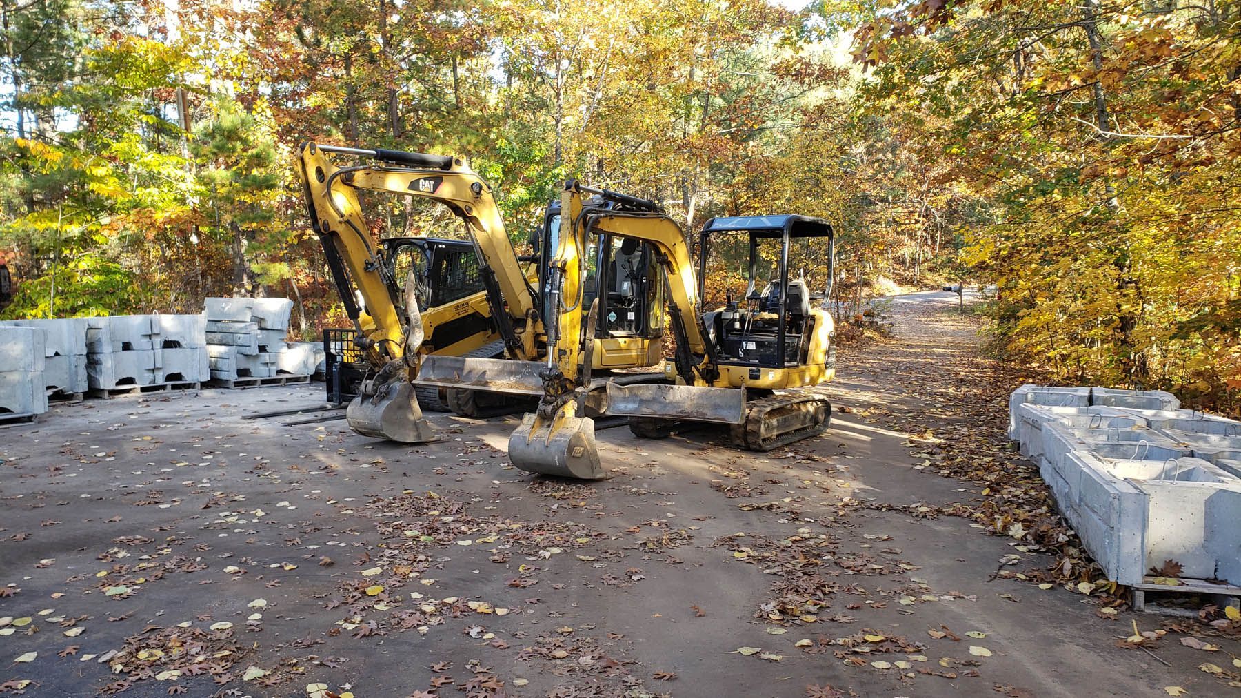 Two yellow excavators parked on a paved area near stacks of gray blocks and trees.