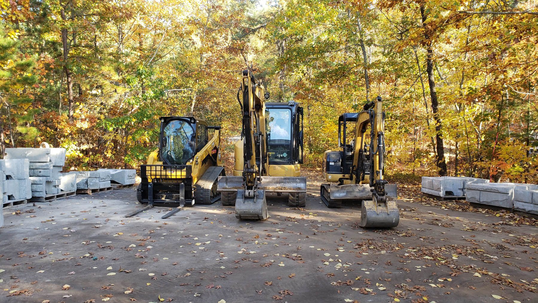 Three yellow excavators parked on pavement in front of trees with autumn foliage.
