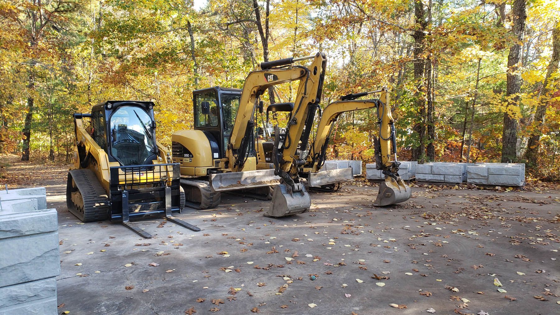 Three yellow construction machines parked outdoors near large gray blocks and fall foliage.
