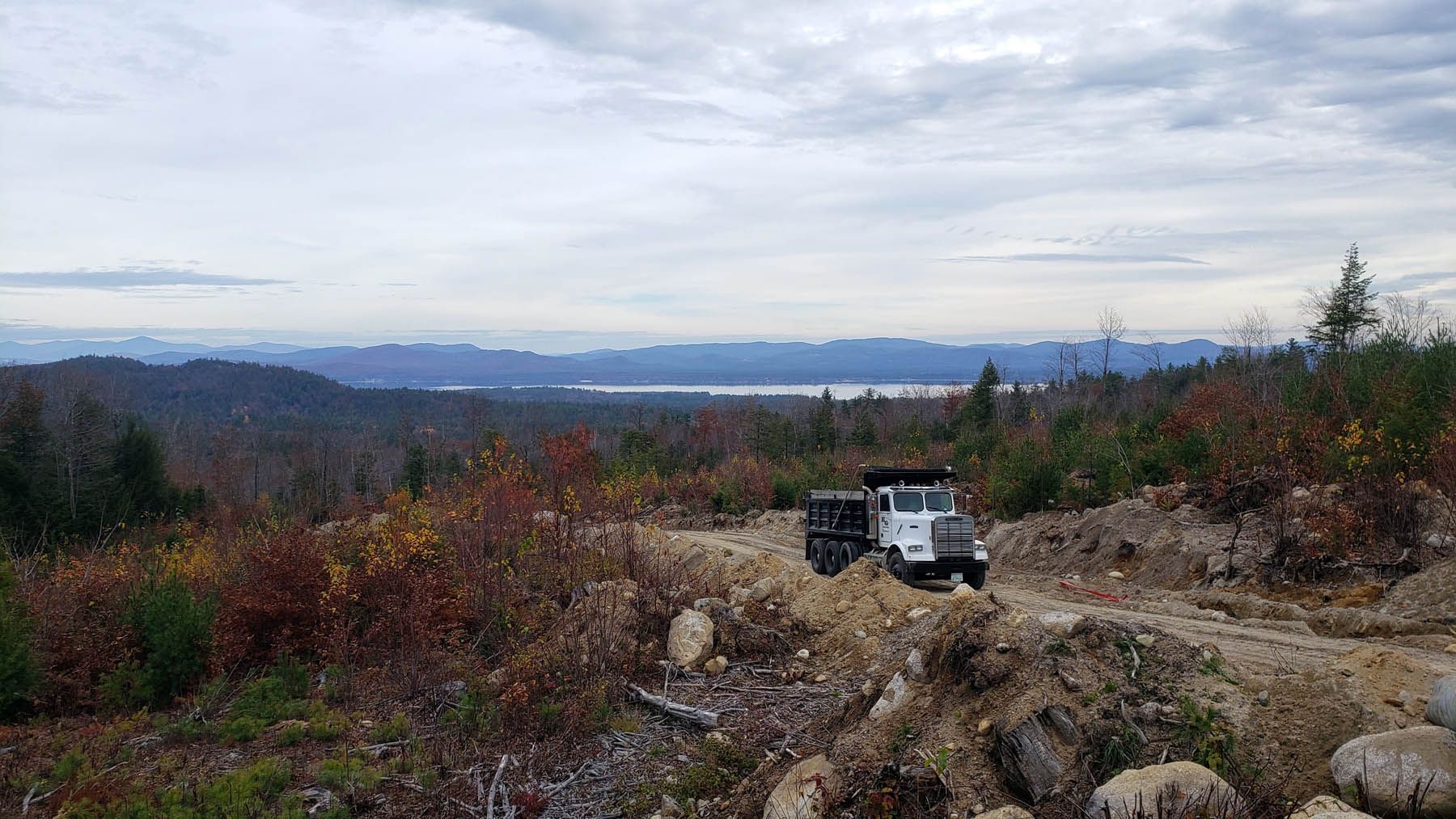Dump truck on a dirt road overlooking a forested landscape and distant lake under a cloudy sky.