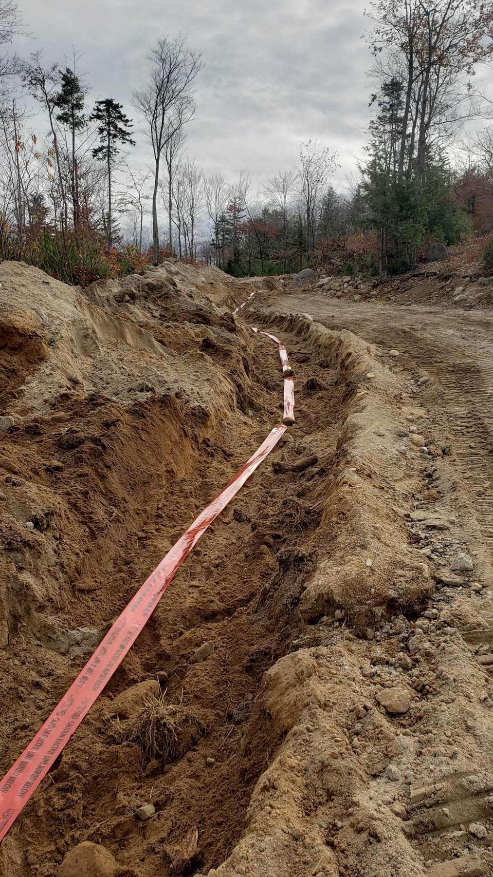 A trench dug in dirt with a red warning tape, possibly for utility lines, in a wooded area.