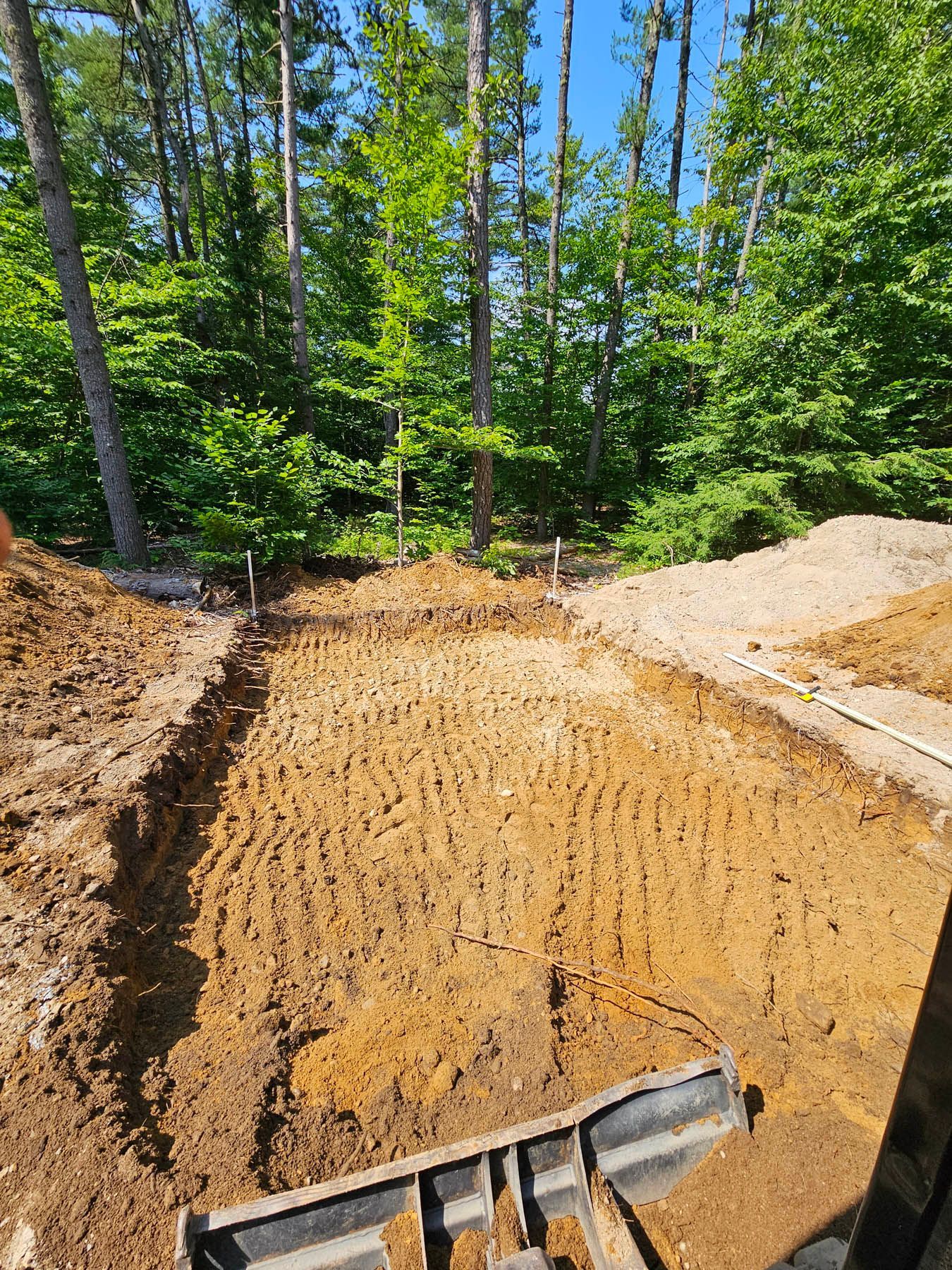 An excavator bucket scoops dirt from a cleared building site in a forest, preparing for construction.