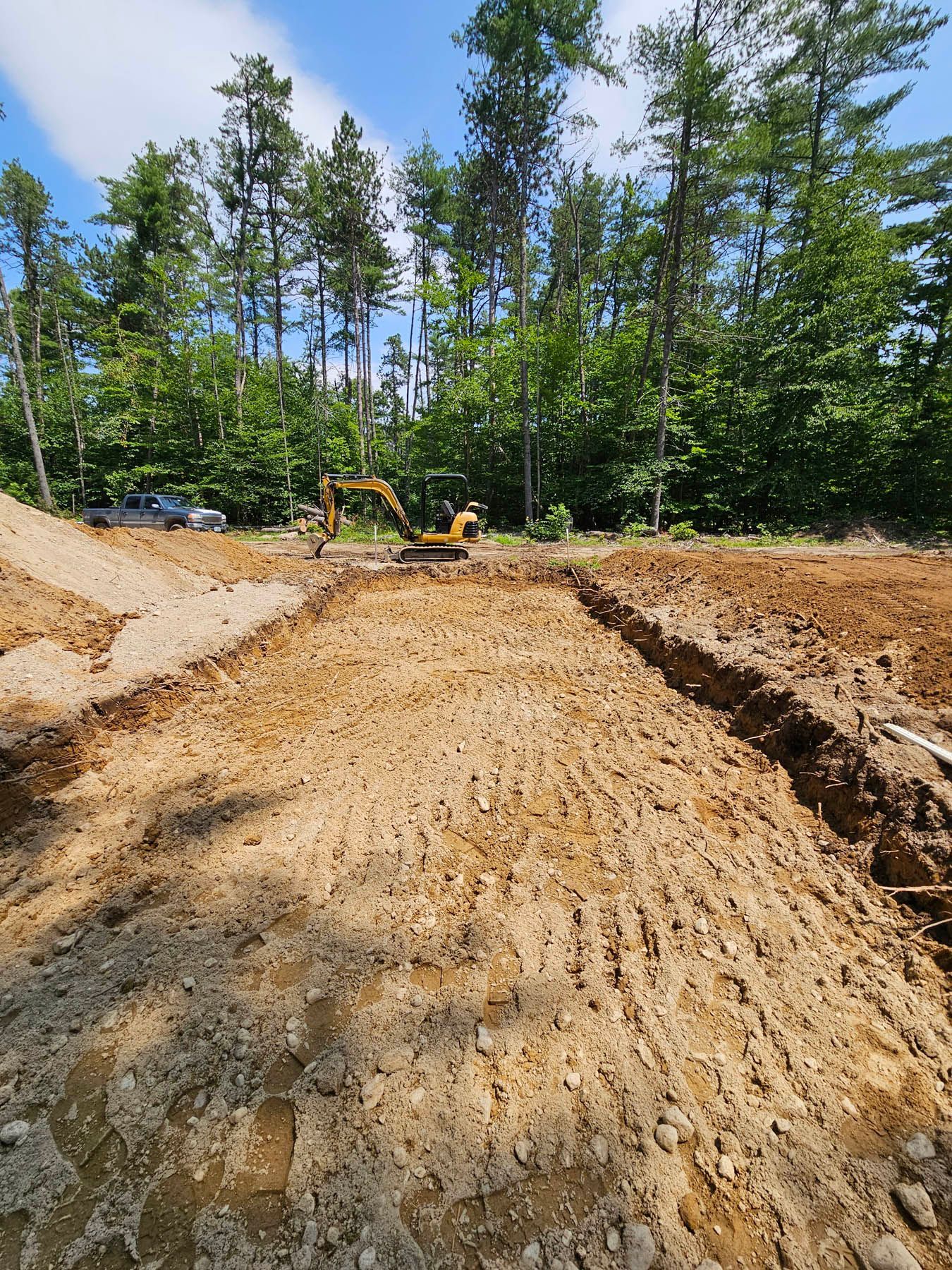 Construction site with an excavator digging trenches in the dirt, trees in the background on a sunny day.
