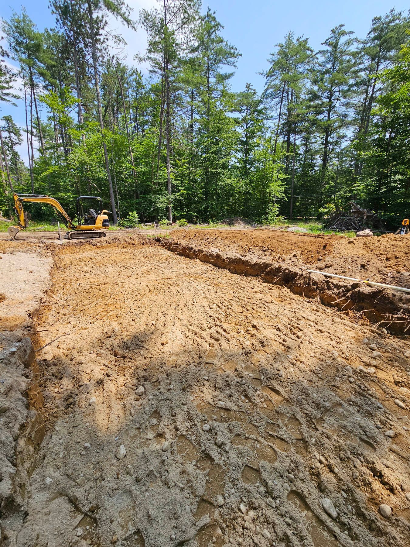 Construction site, excavator digging a trench in brown soil, trees in the background under a blue sky.