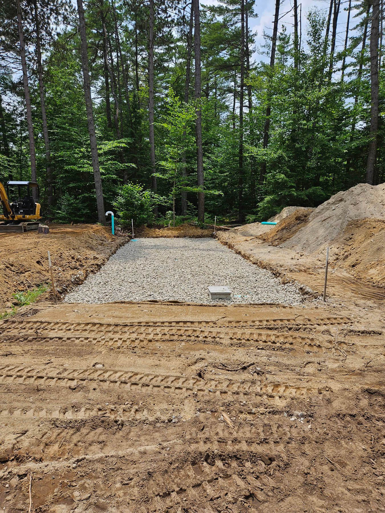 Gravel-filled rectangular pit in a dirt clearing, possibly for a septic system, surrounded by trees and construction equipment.