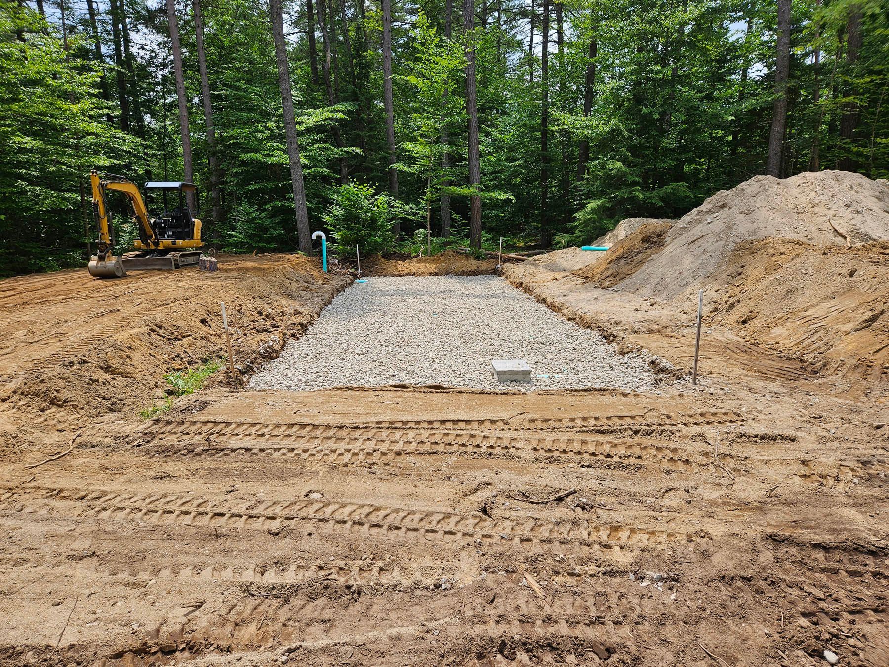 Construction site with an excavator and gravel-filled rectangular foundation, surrounded by dirt and trees.