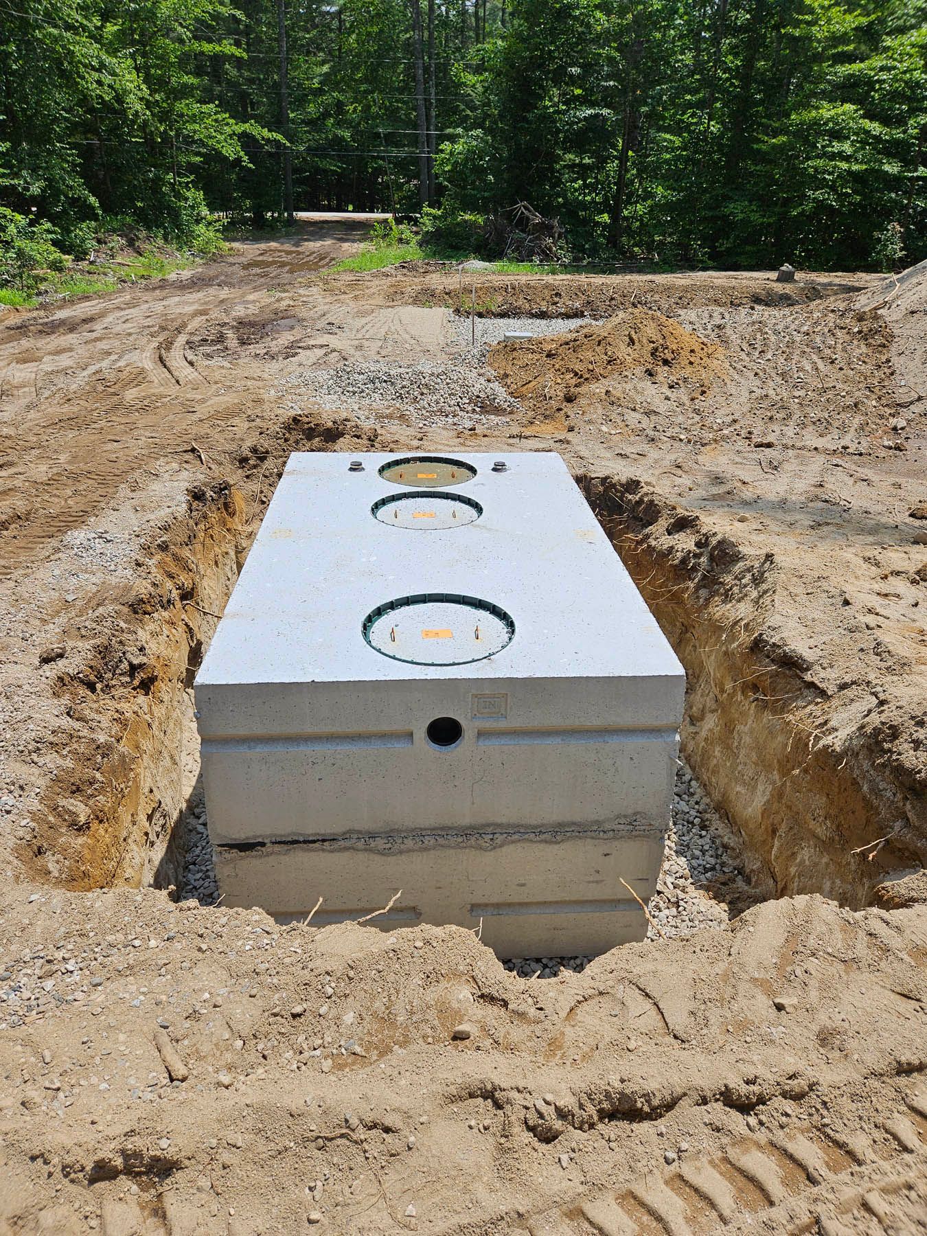 Concrete septic tank installed in a dirt trench, ready for backfill, in a wooded area.