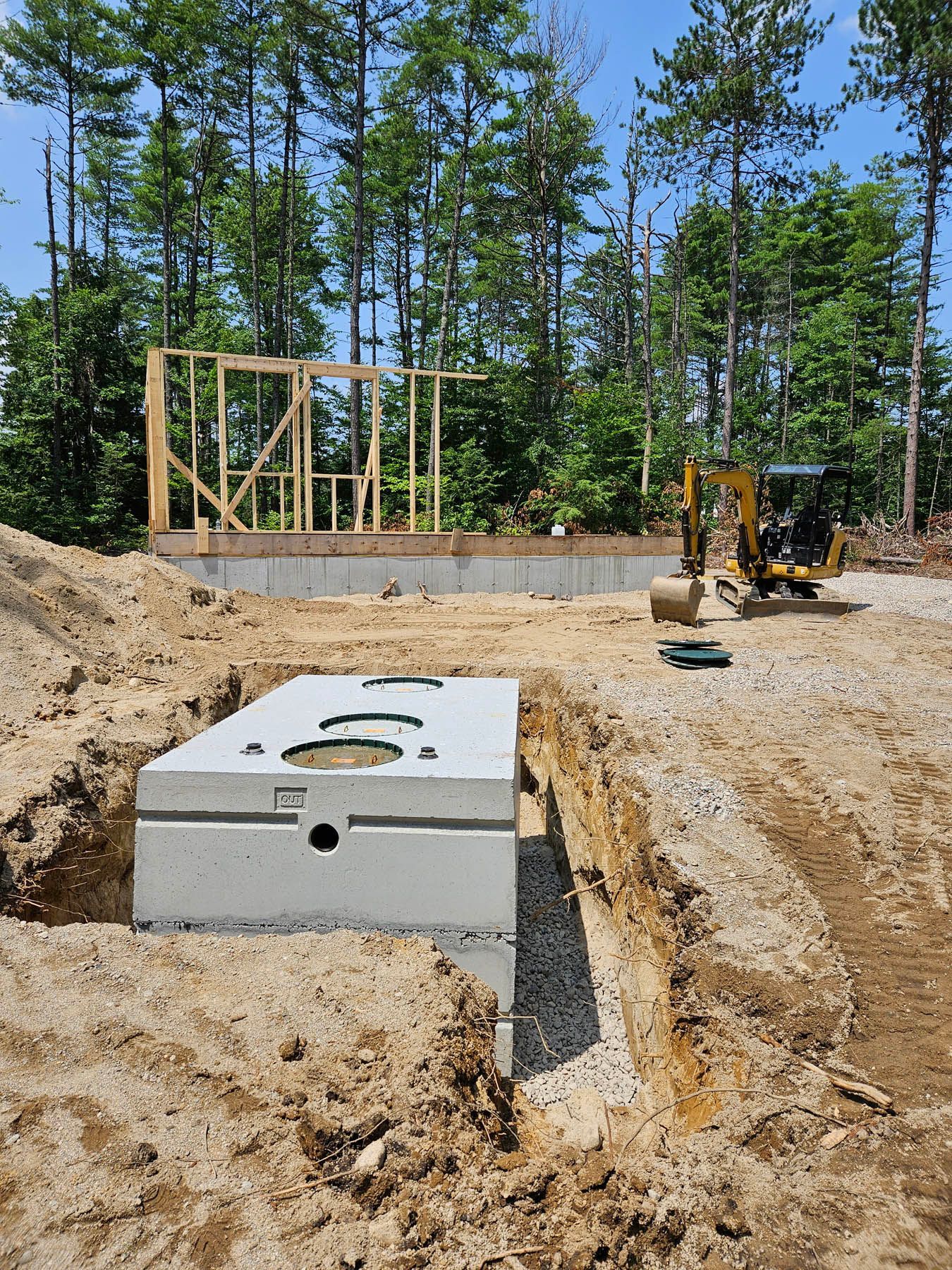 A concrete septic tank in an excavated trench, with a new house under construction in the background.