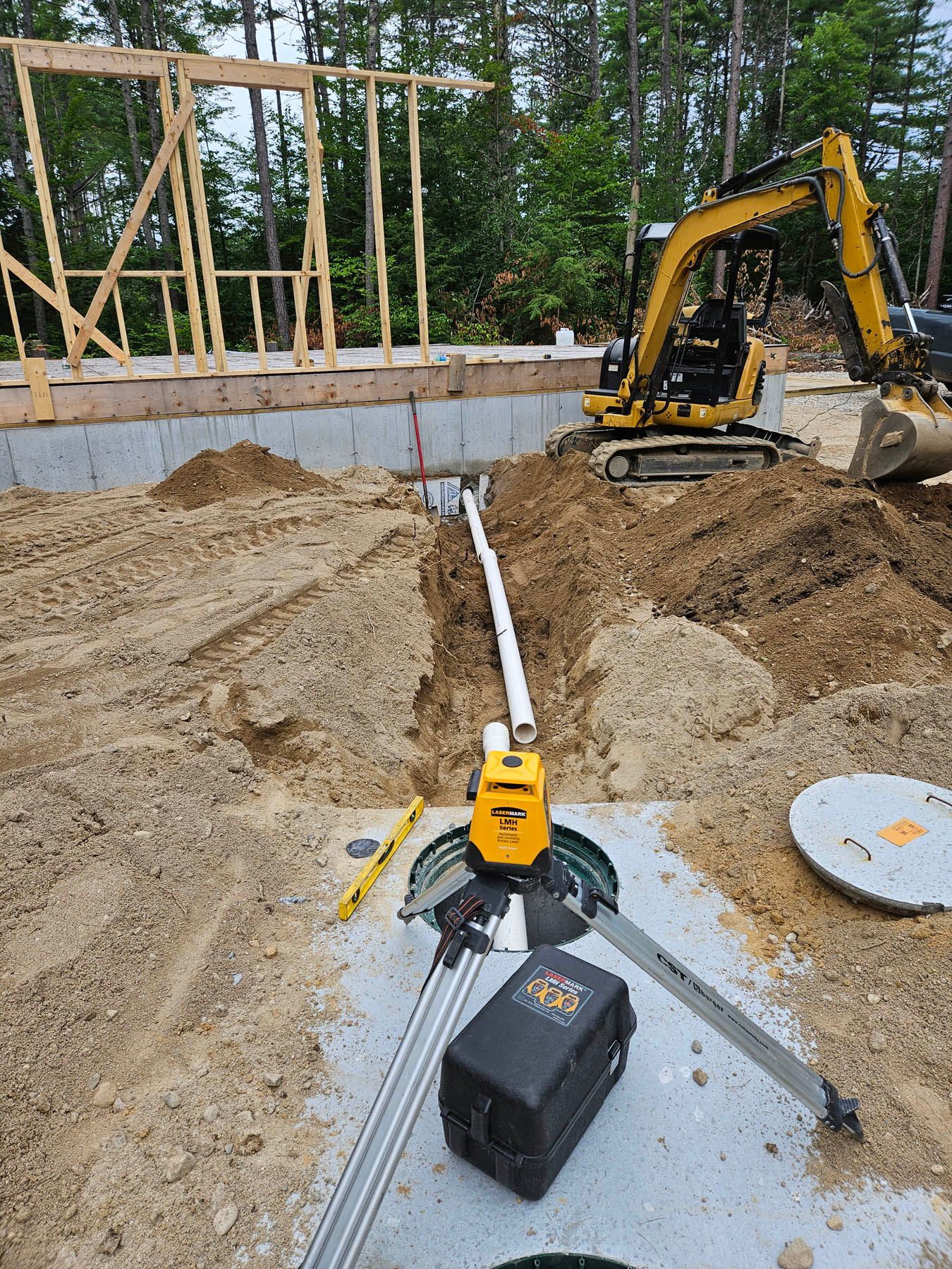 Construction site: Excavator, laser level, and pipes in trench, near a foundation and wood framing.