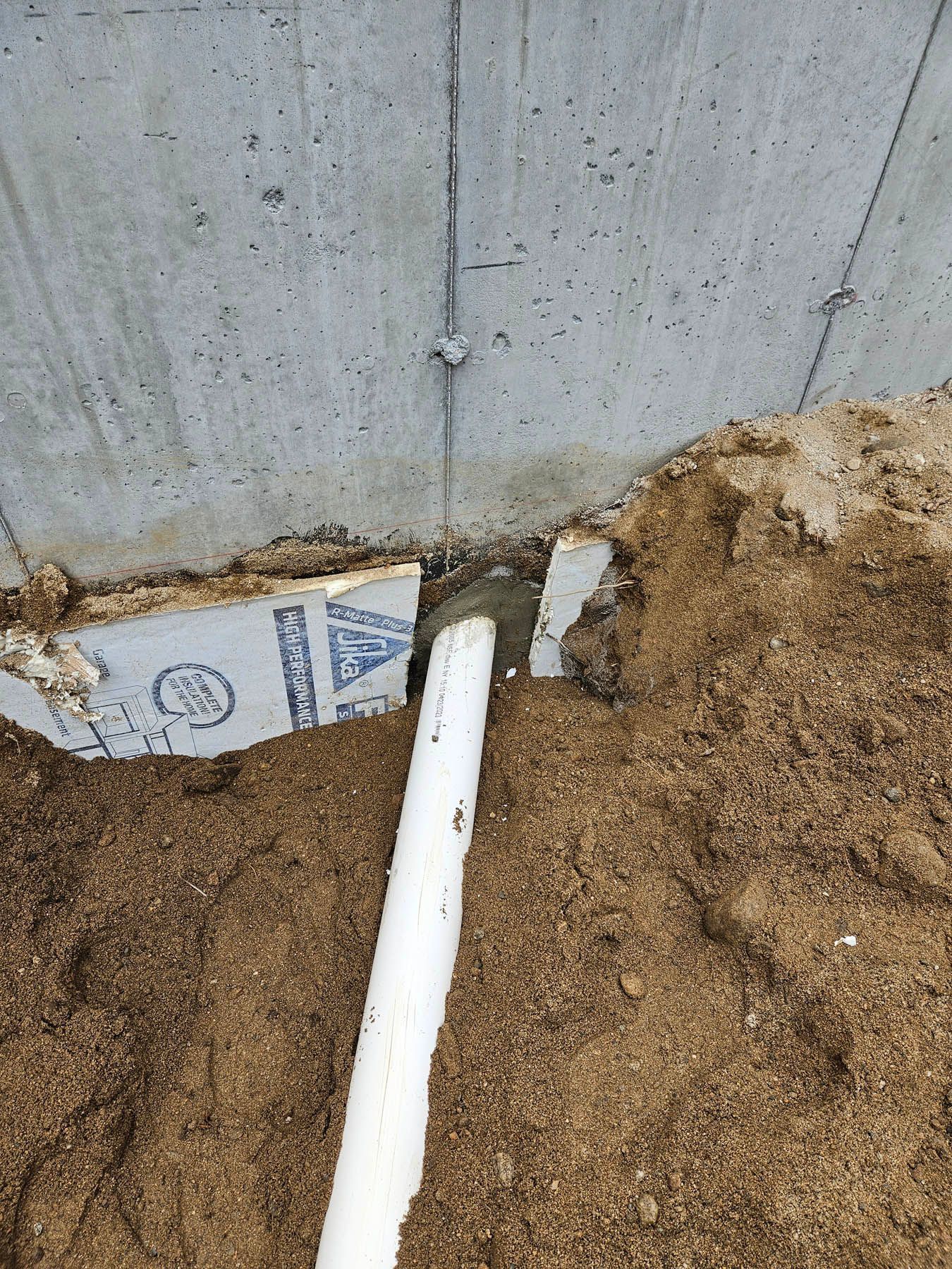 White PVC pipe extending from a concrete foundation, surrounded by dirt and insulation.