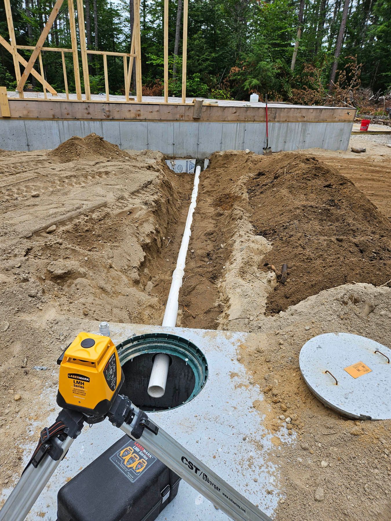 Construction site: a white pipe in a trench, leading to a tank. Yellow laser level on a tripod, dirt piles, foundation.