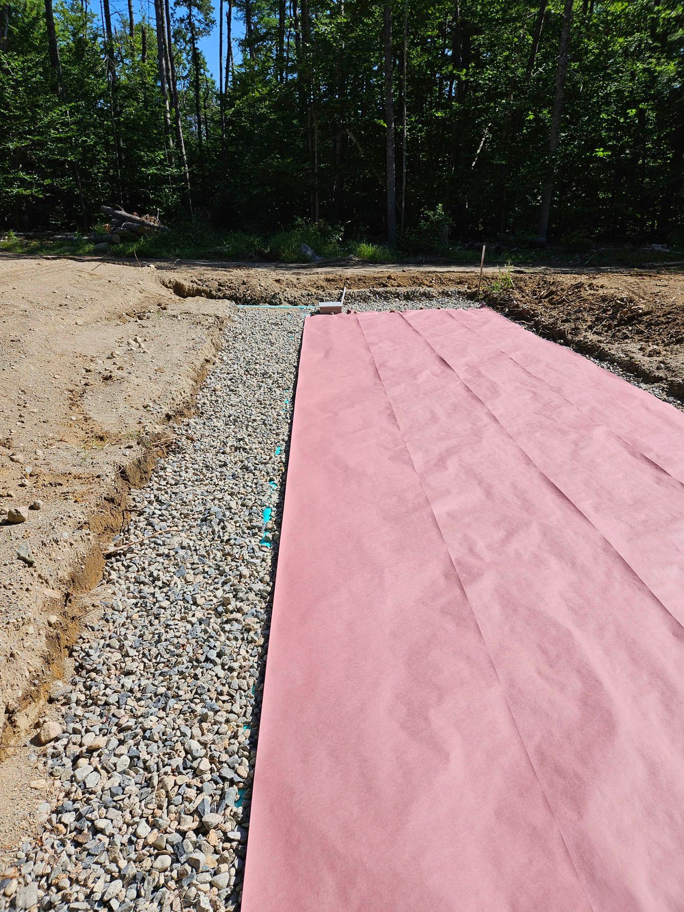 Pink landscape fabric laid over gravel in a trench, near a dirt path and forest.