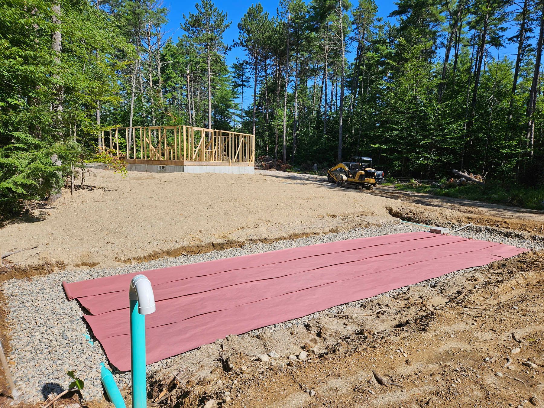 Construction site with a house foundation, gravel, and pink ground cover in a wooded area.