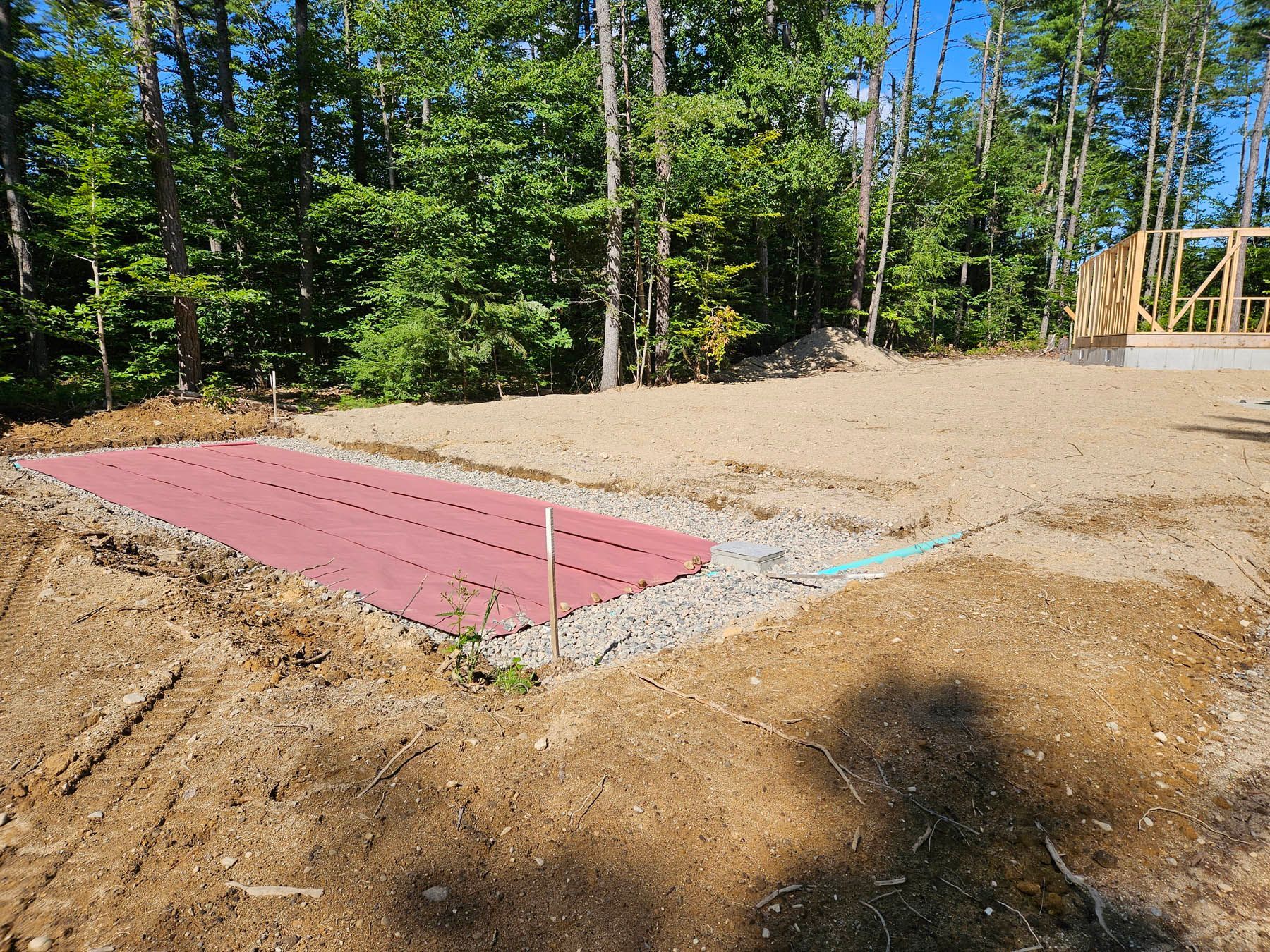A construction site with a red septic field. Dirt and gravel surround the field and a frame of a building in the background.