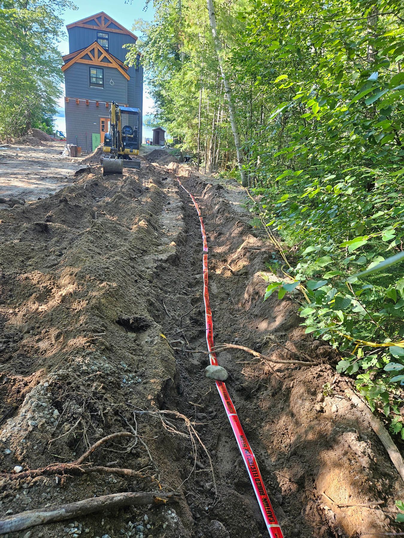 Red conduit in a trench being laid near a blue house and trees; construction site.