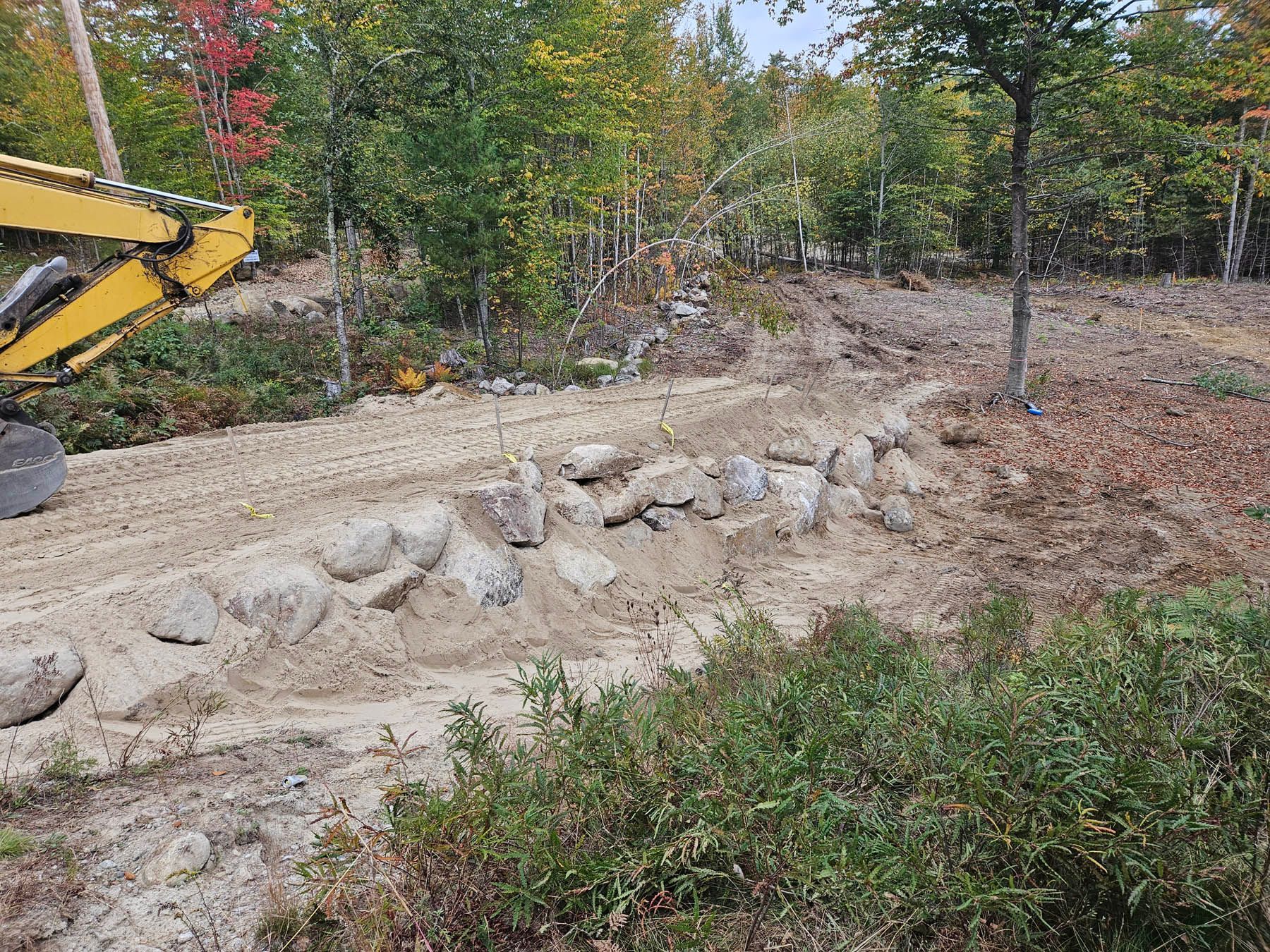 Construction site with a rock wall, dirt, and yellow excavator against a forested backdrop.