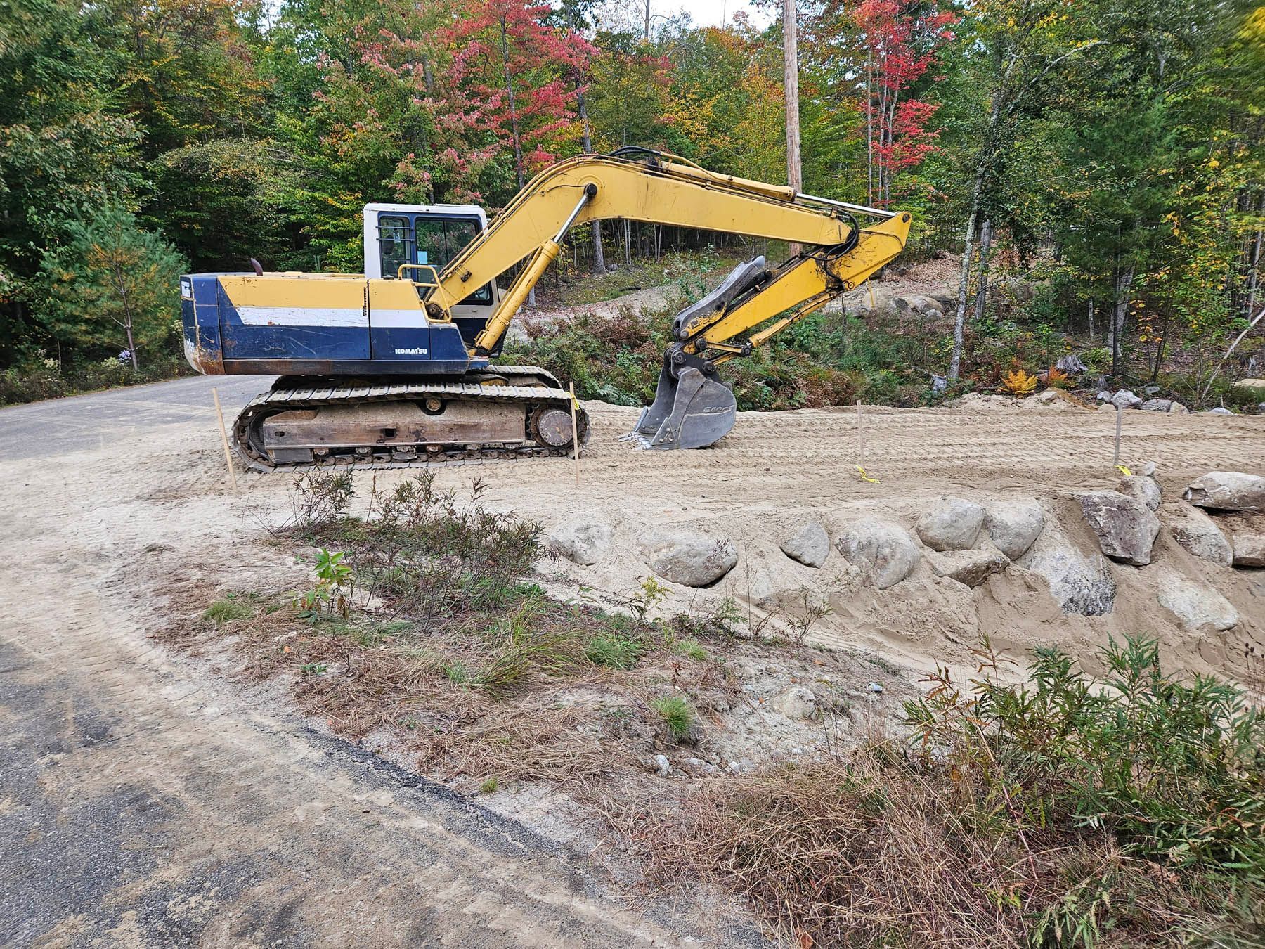 Yellow and blue excavator on a gravel surface, with trees in the background.