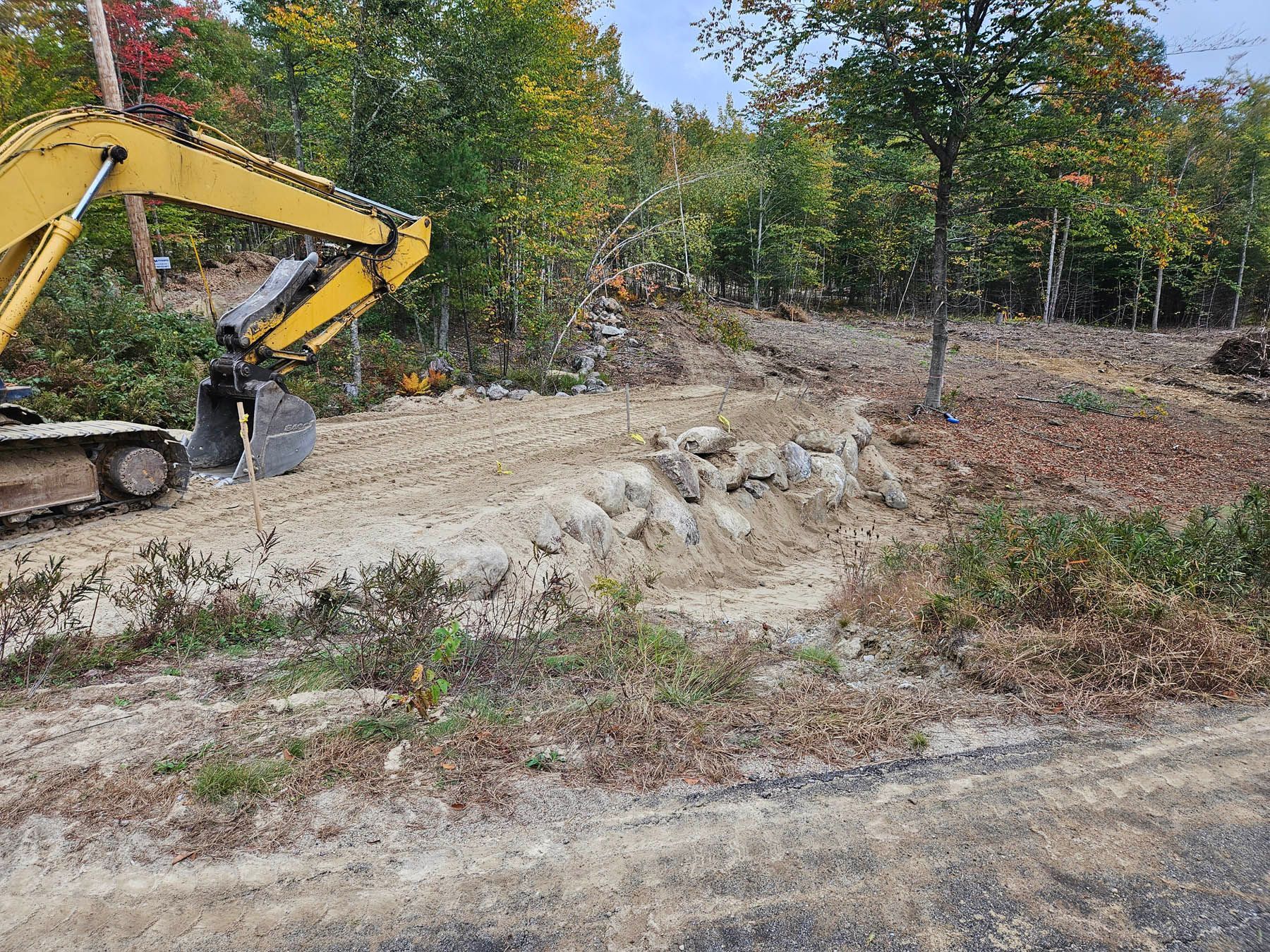 Yellow excavator on a dirt clearing next to a rock wall, trees, and brush under a blue sky.