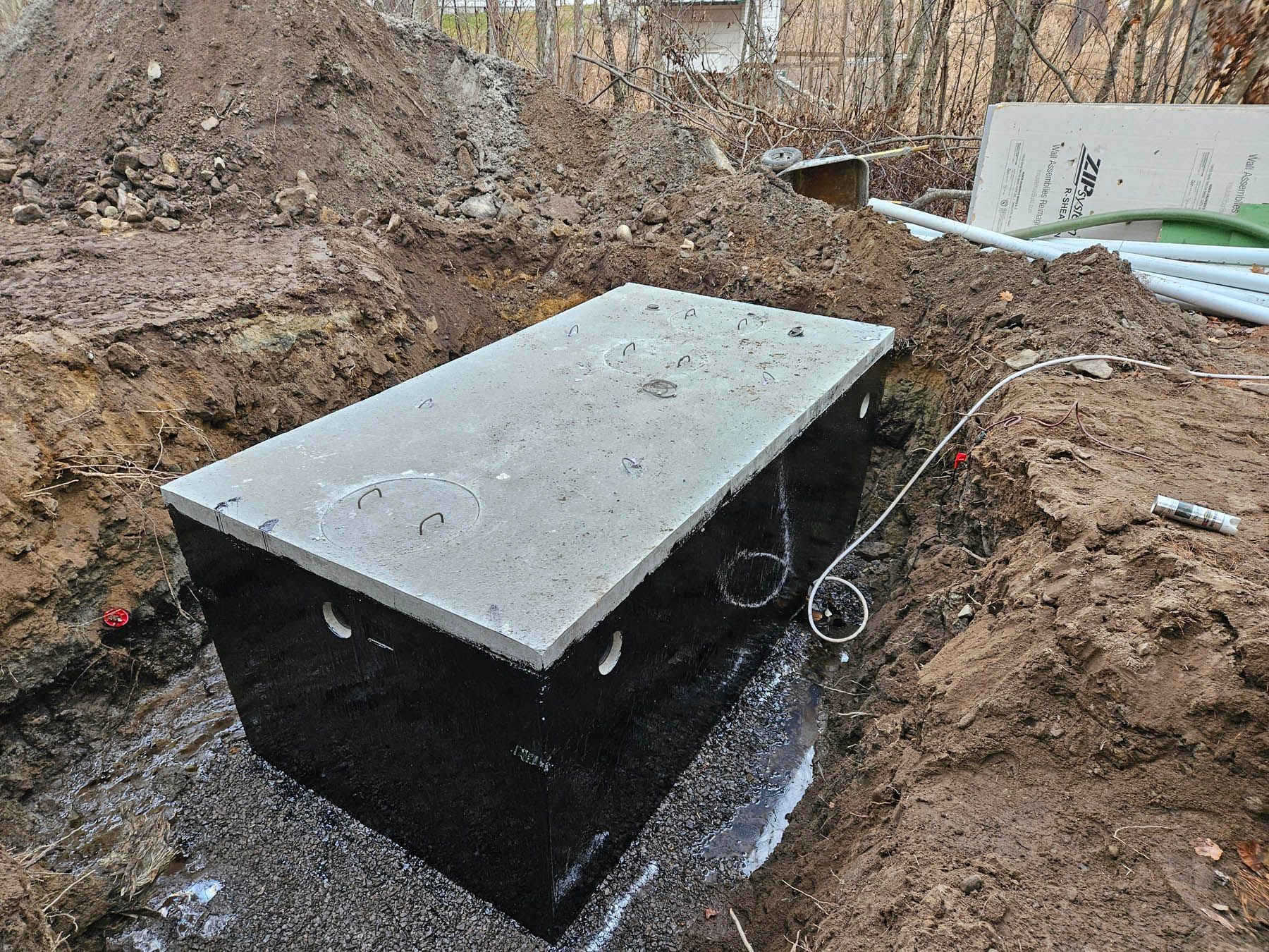 Rectangular septic tank in a dirt excavation. Black sides, concrete top. Pipes visible.