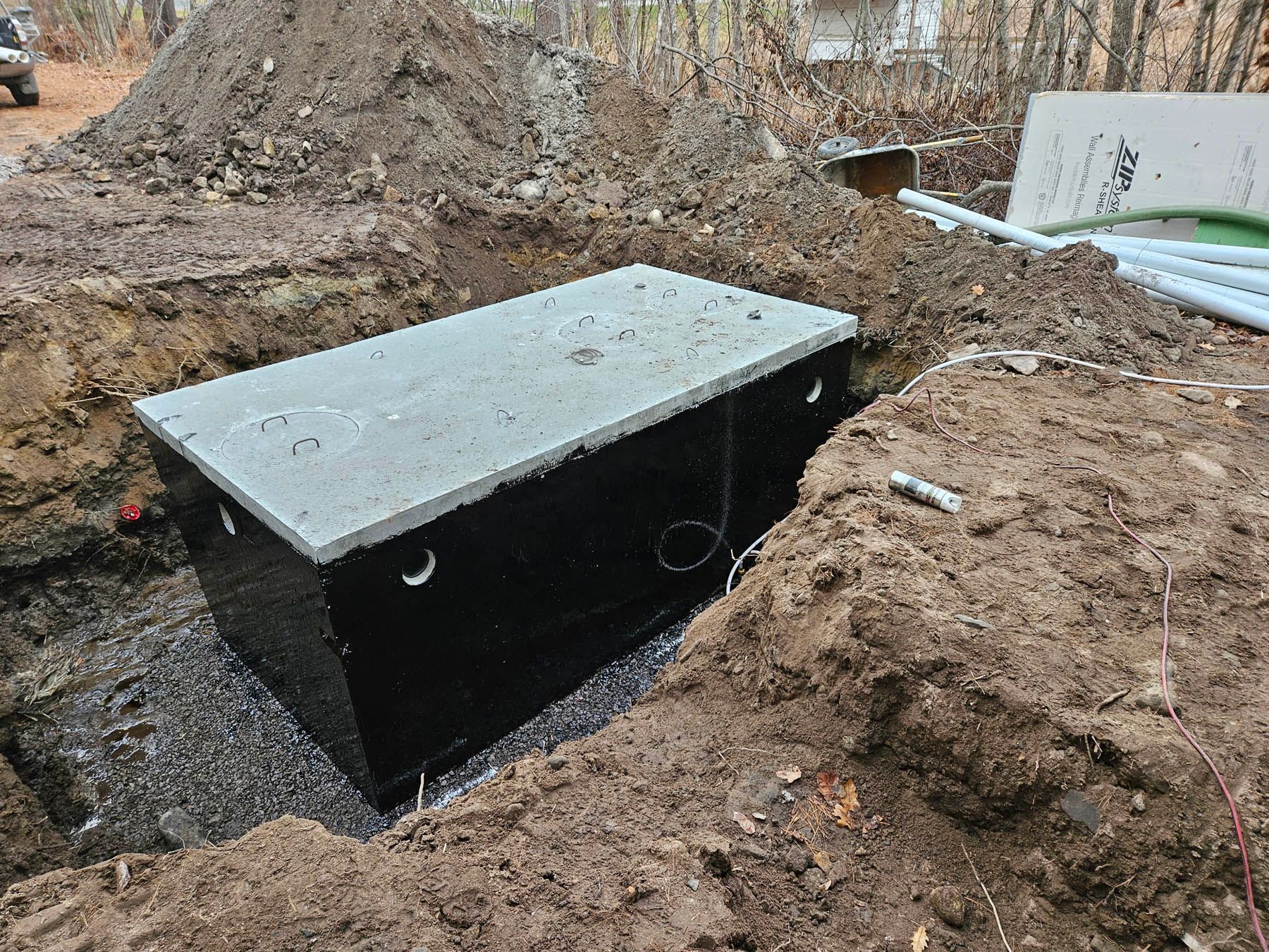 A rectangular concrete septic tank in a trench, with a dark coating, in an outdoor setting.