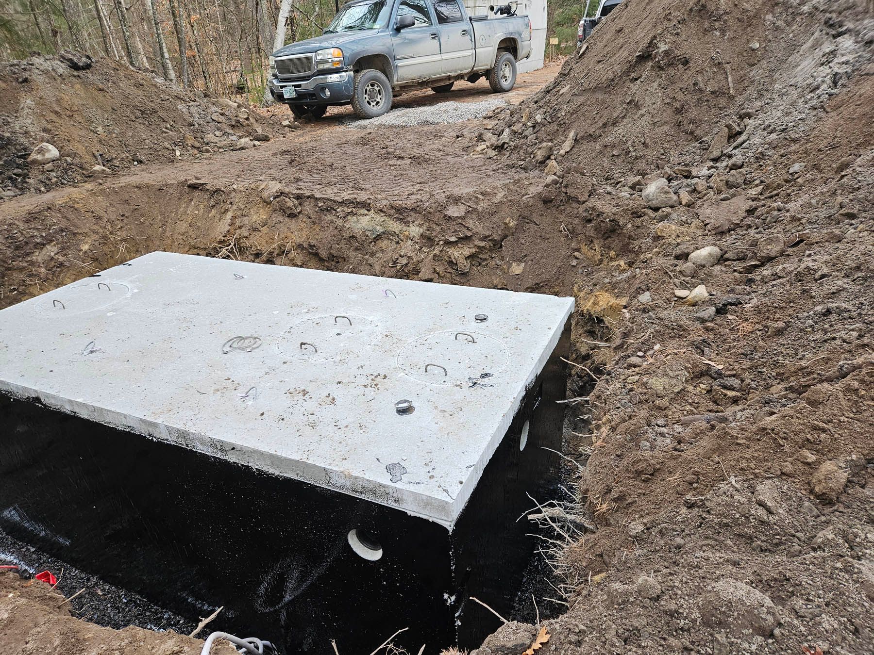 Concrete septic tank partially buried in a dirt trench, truck in the background.