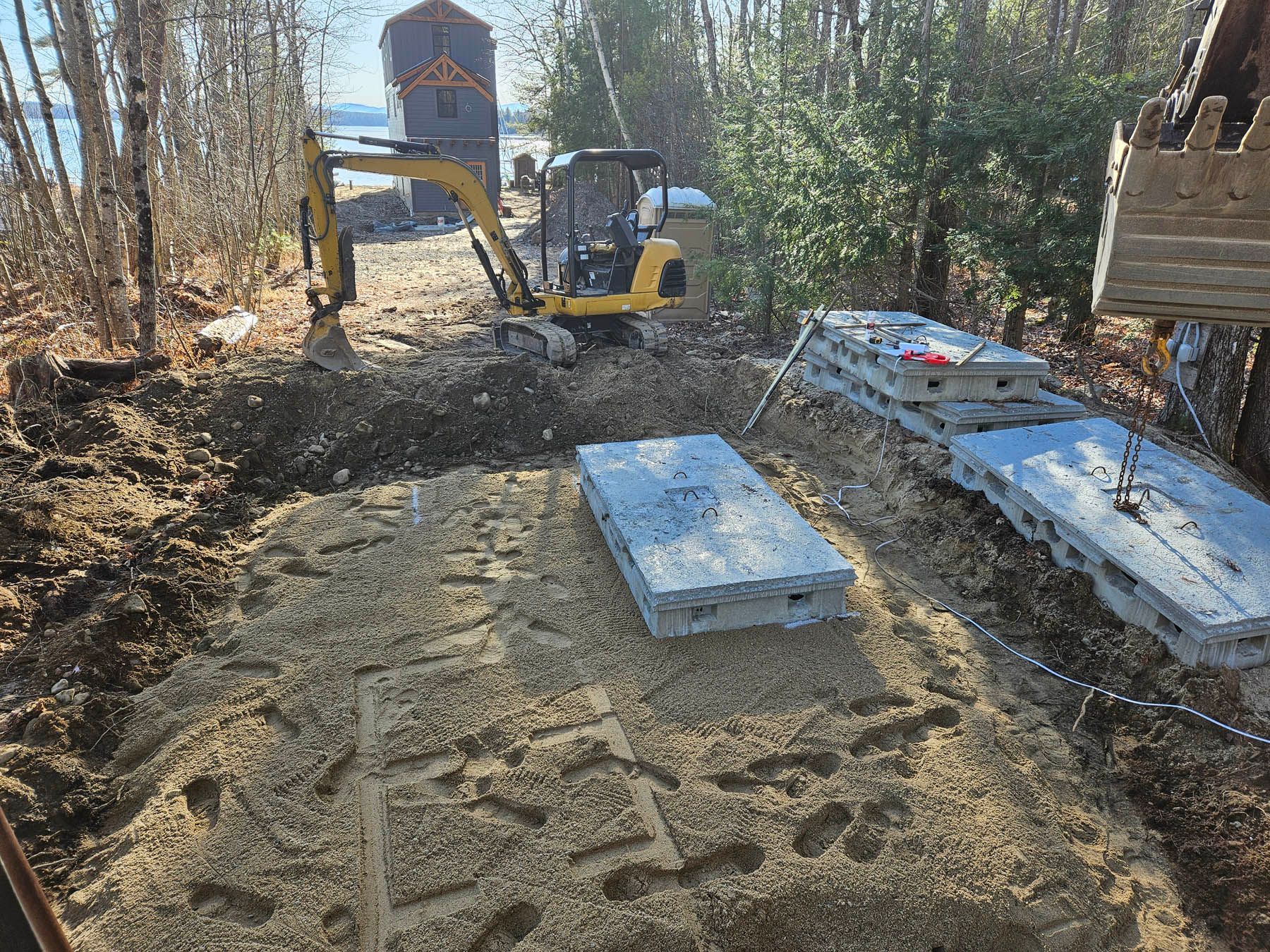 Excavator digging, concrete slabs ready. Construction site near water with trees.