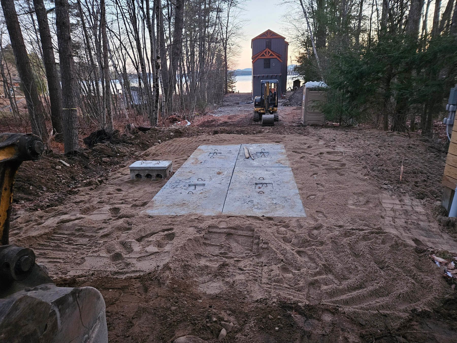 A concrete septic tank cover surrounded by dirt, trees, and a small excavator in a clearing.