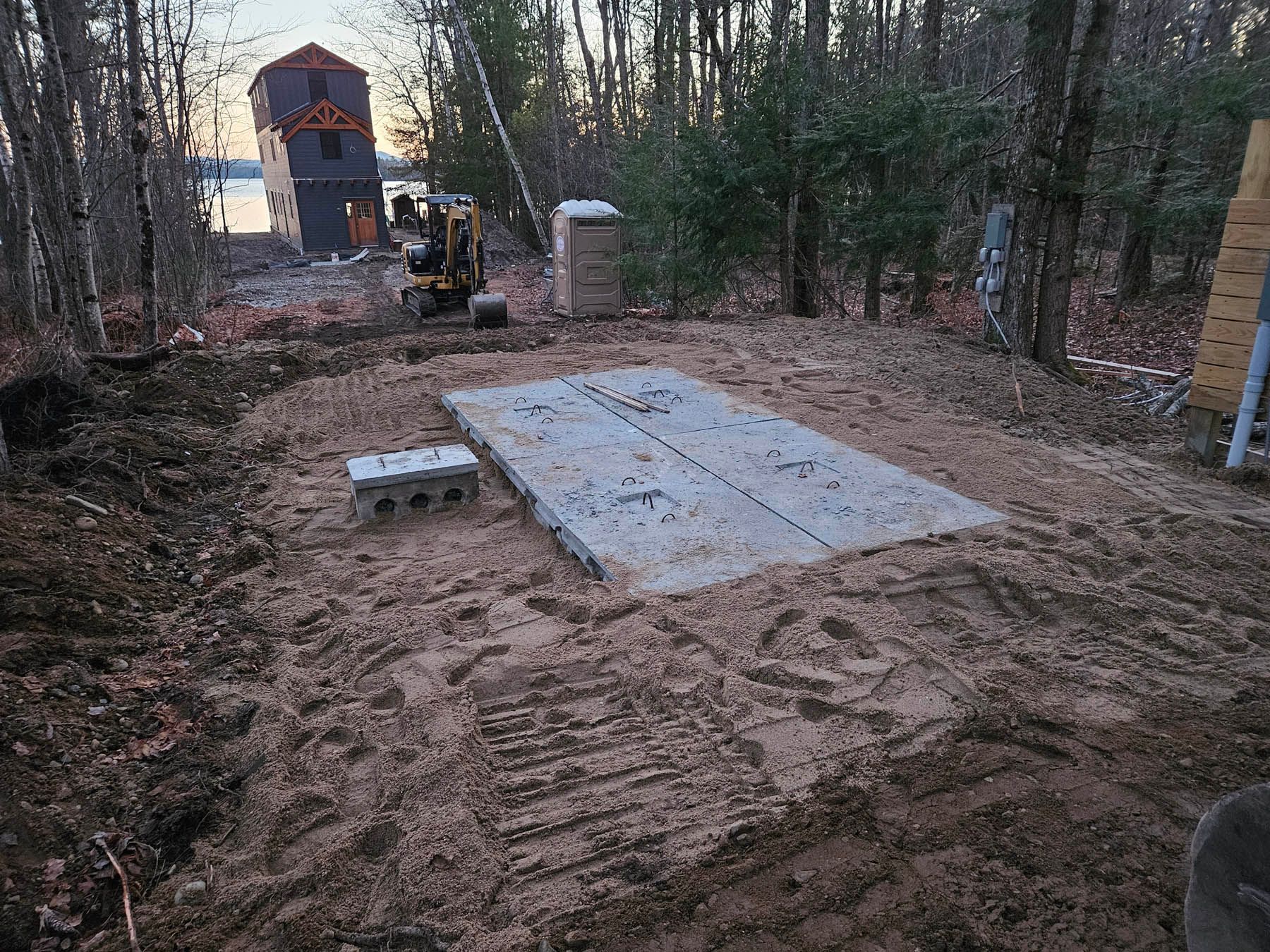 Concrete slab in a sandy clearing with excavator, near trees, and a building under construction.