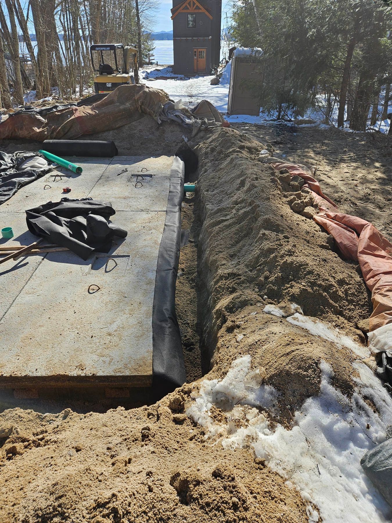 Excavation site with a trench, concrete slab, and small excavator. Snow present. Outdoors, sunny day.