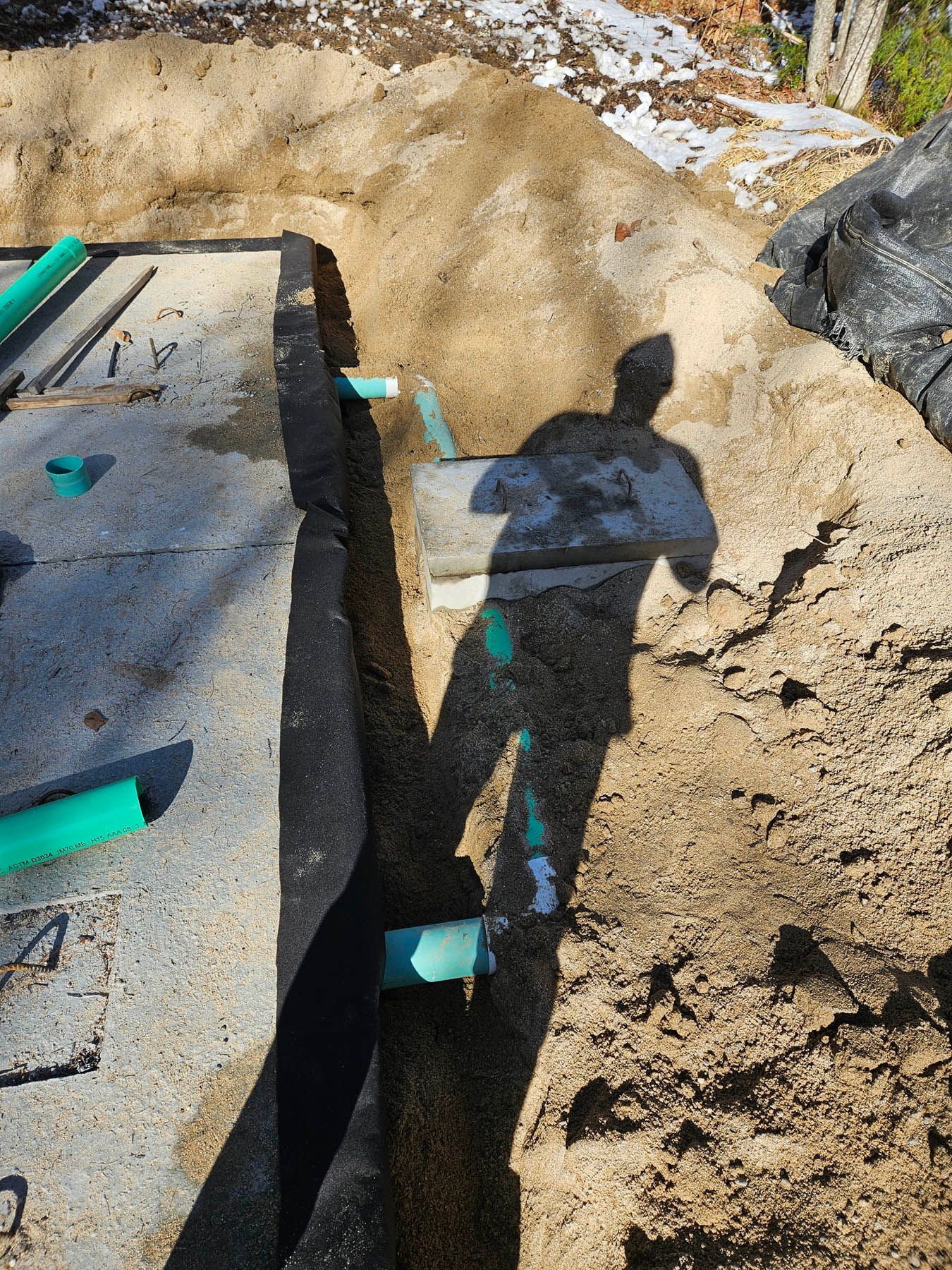 A construction site with green pipes emerging from the ground, covered by dark fabric and a person's shadow.