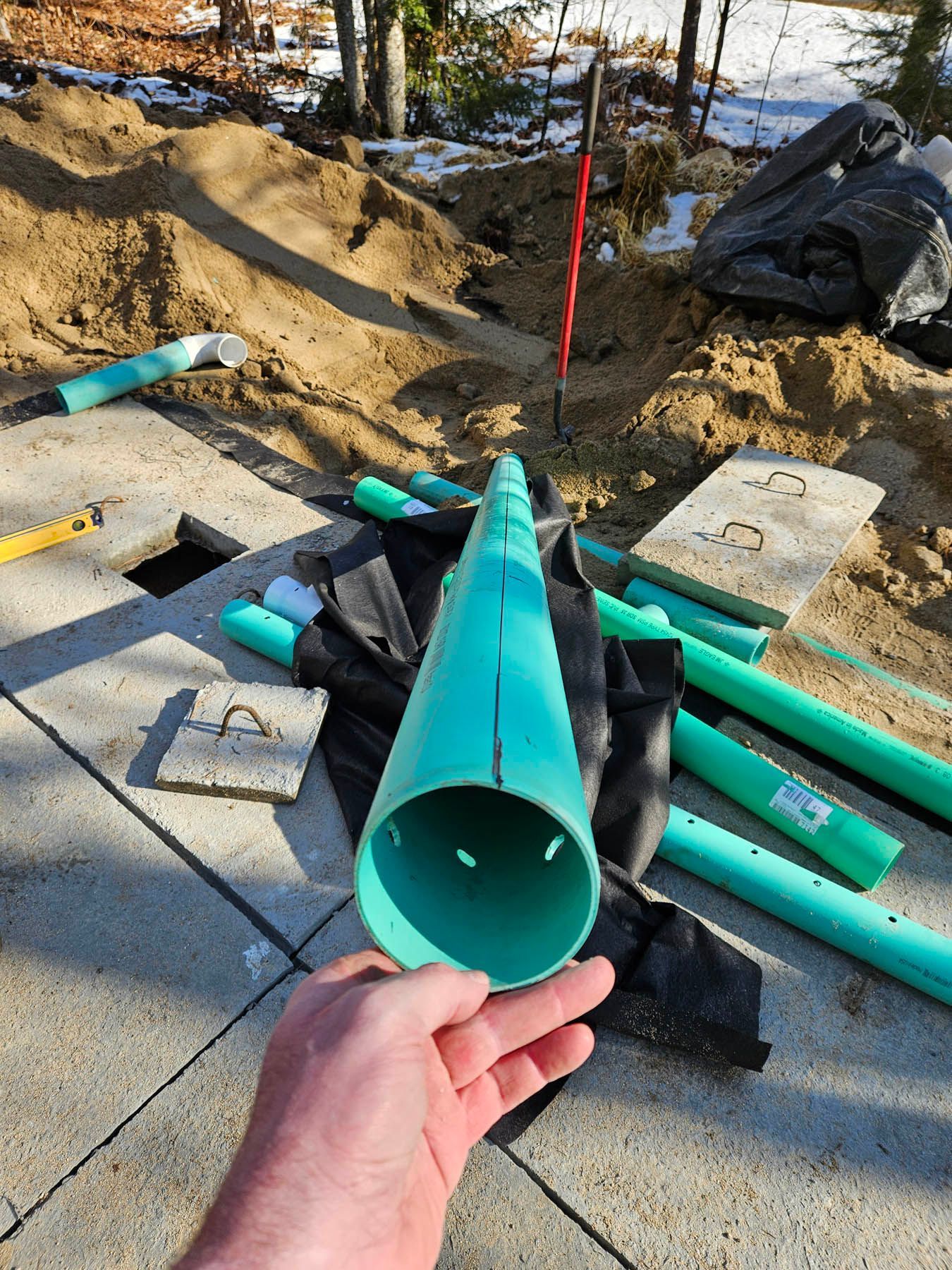 Hand holding a green PVC pipe, with other pipes and construction materials in a yard.