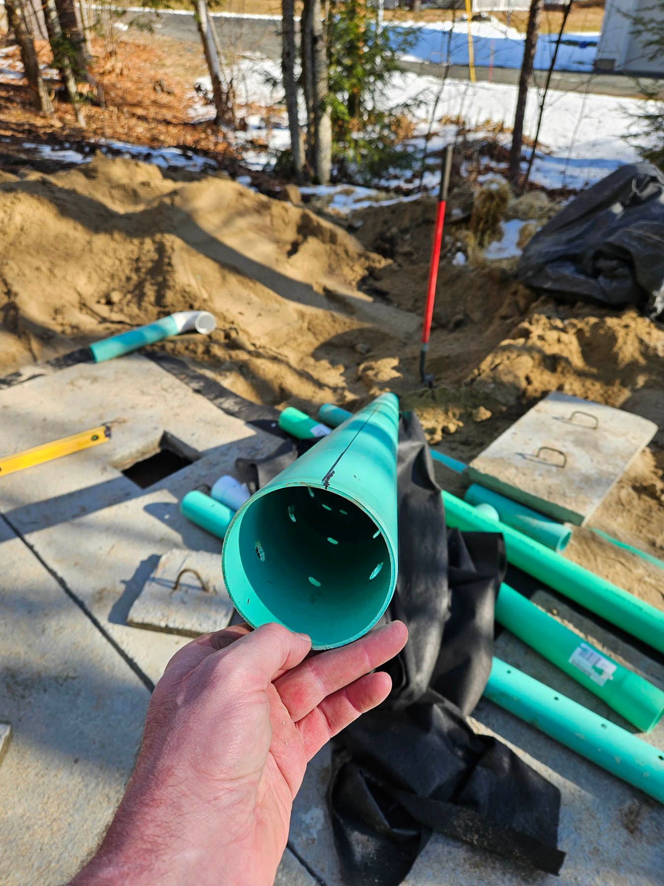 Hand holding a perforated green pipe, construction site with trench, pipes, and tools.