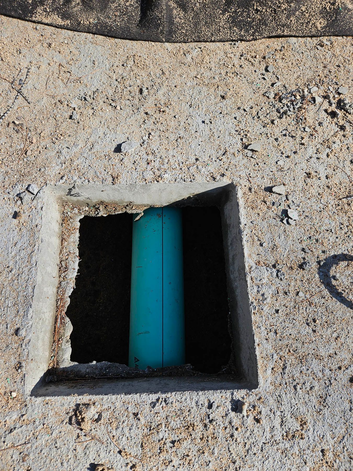 Blue PVC pipe visible through a square hole cut in a rough, light-colored concrete surface.