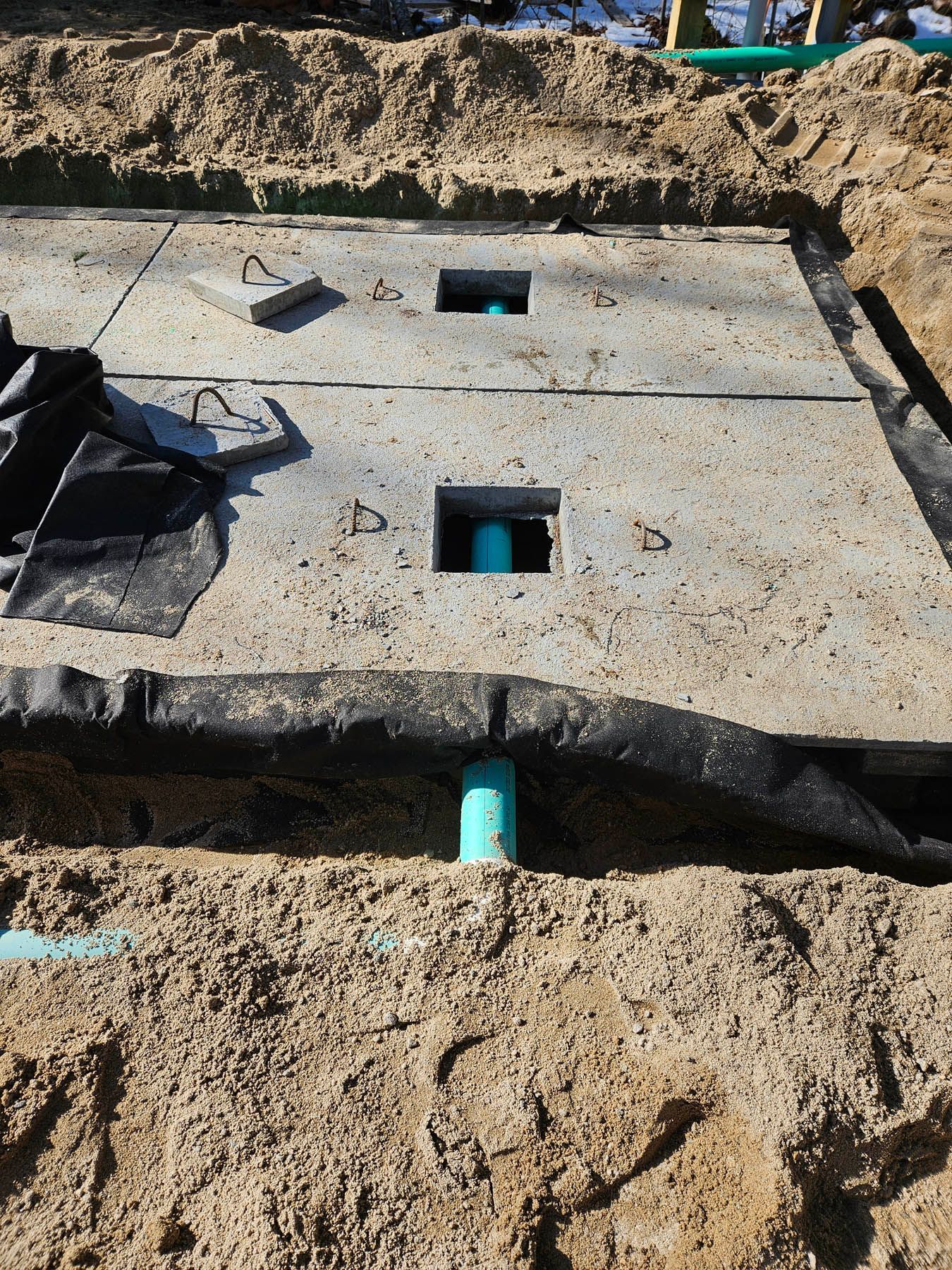 Concrete septic tank lid with access holes, set in sandy soil. Blue pipes are visible.